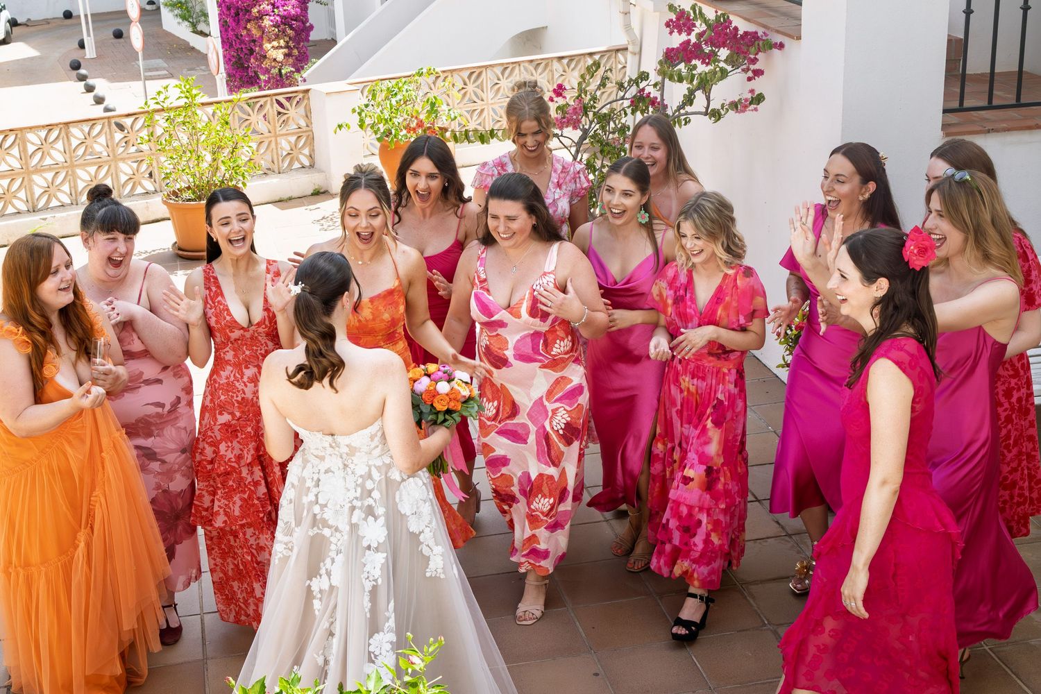 Bride in floral gown surprises bridesmaids in vibrant pink and orange dresses on a sunlit terrace.