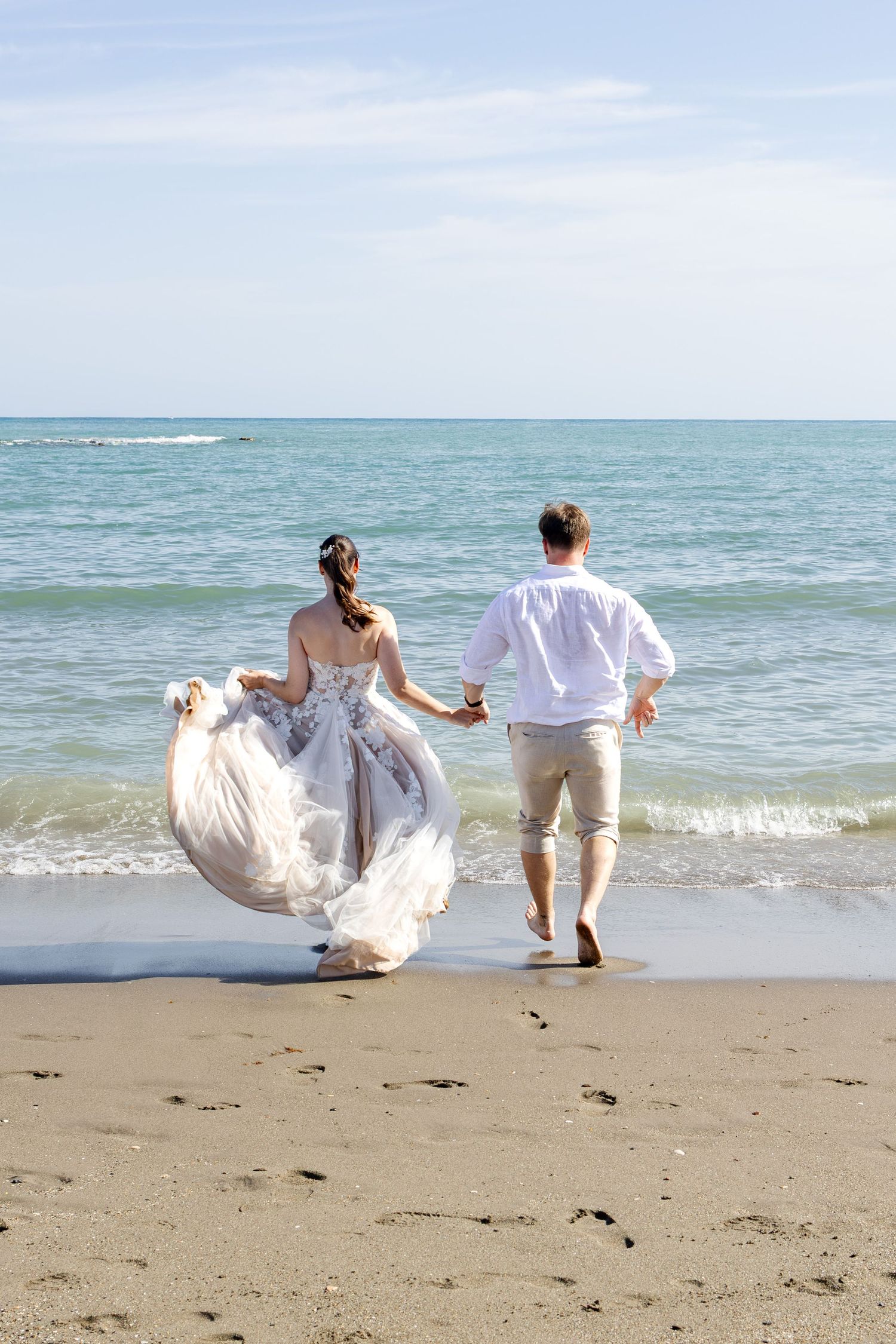 A bride and groom run hand in hand toward the ocean waves on a sunny beach after their wedding.