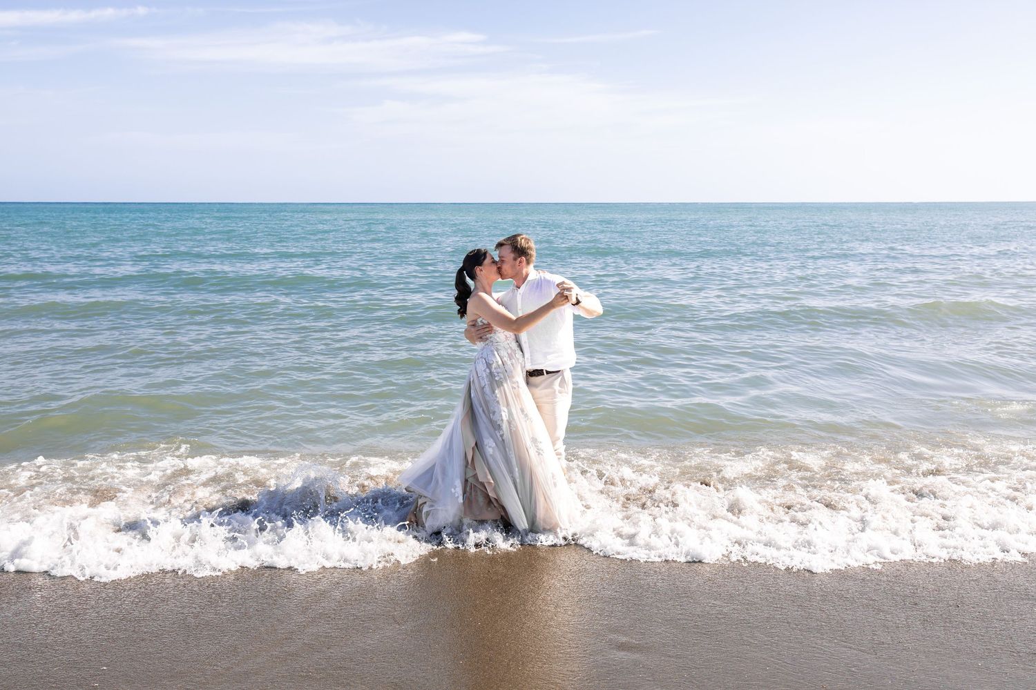 Couple in wedding attire embracing on a sandy beach with ocean waves, romantic destination wedding photo.