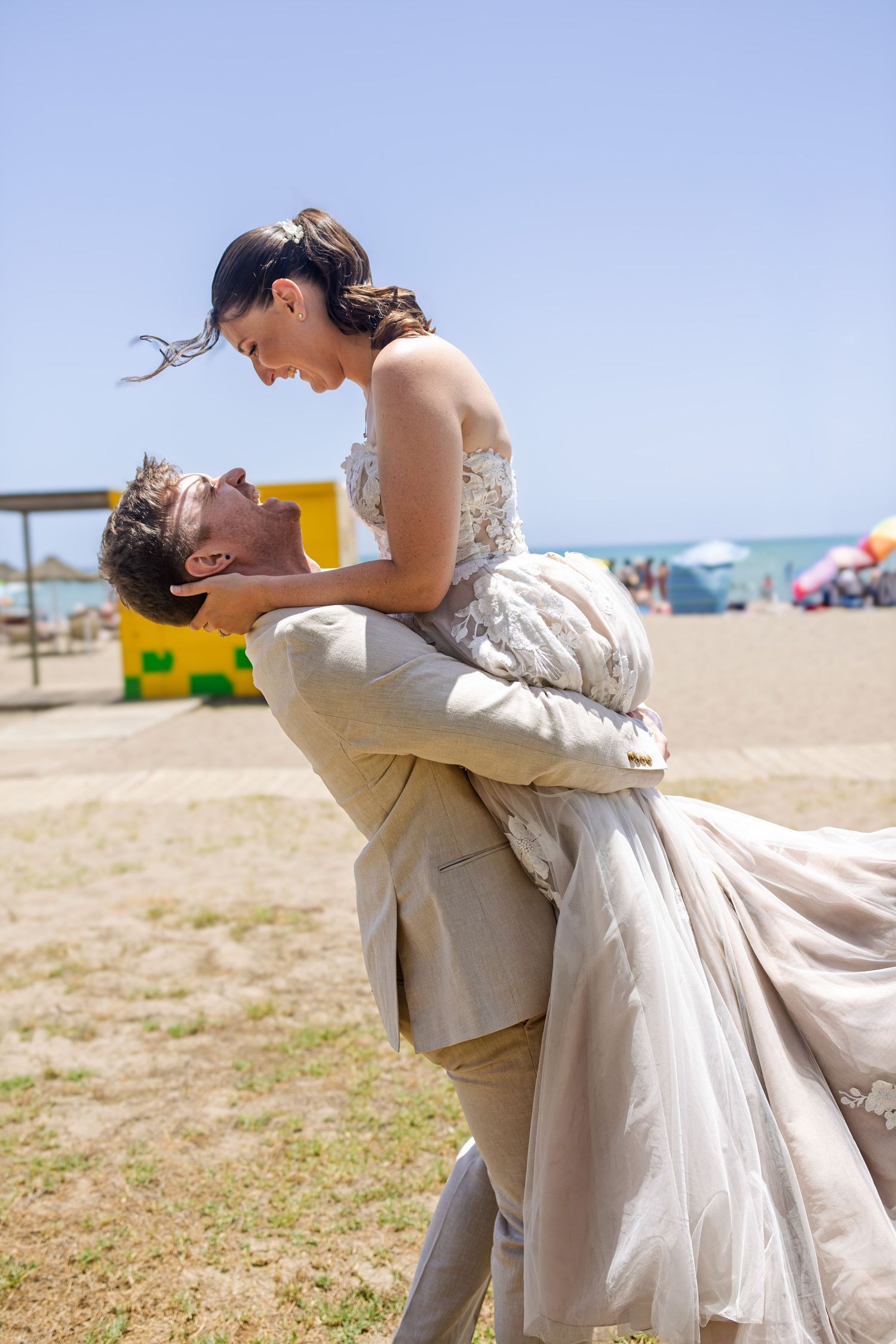 Groom lifting bride in wedding dress on a sunny beach, sharing a romantic moment together.