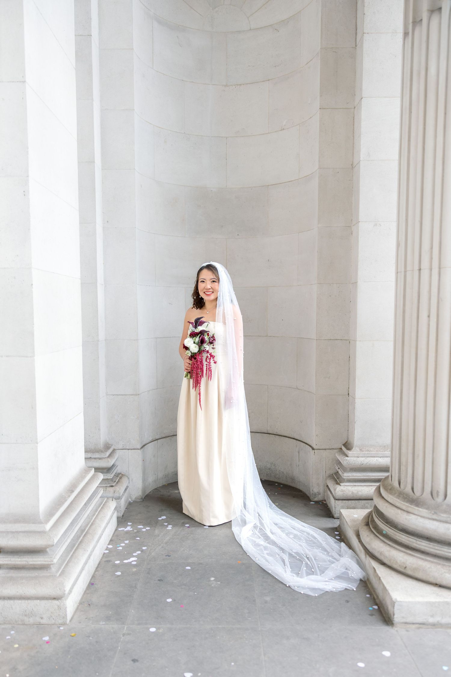 Elegant bride in ivory gown with long cathedral veil holding bouquet between grand columns.