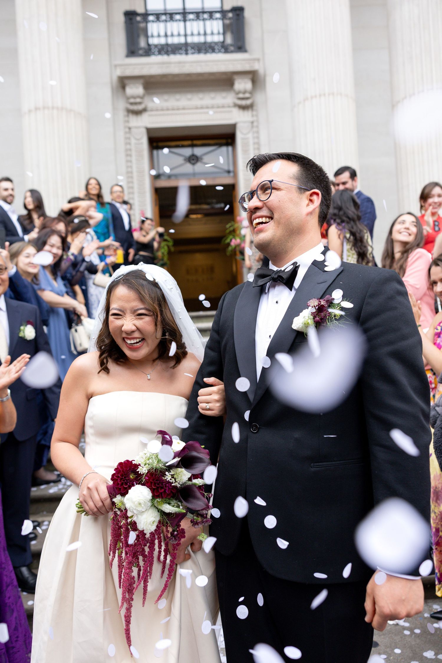 Joyful newlyweds exit grand building as guests shower them with white confetti during wedding celebration.