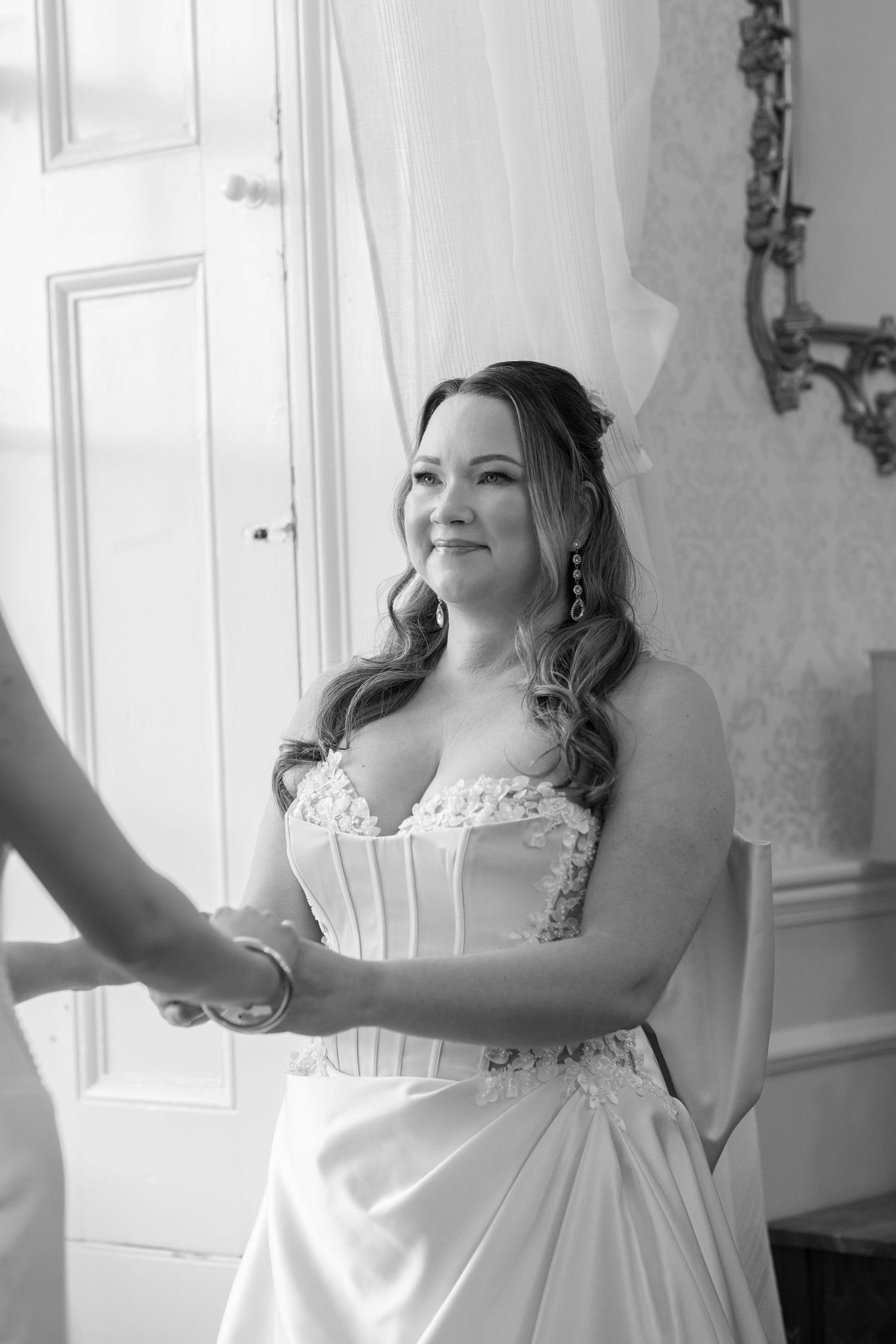 Black and white portrait of someone in an ornate wedding dress standing by a window.