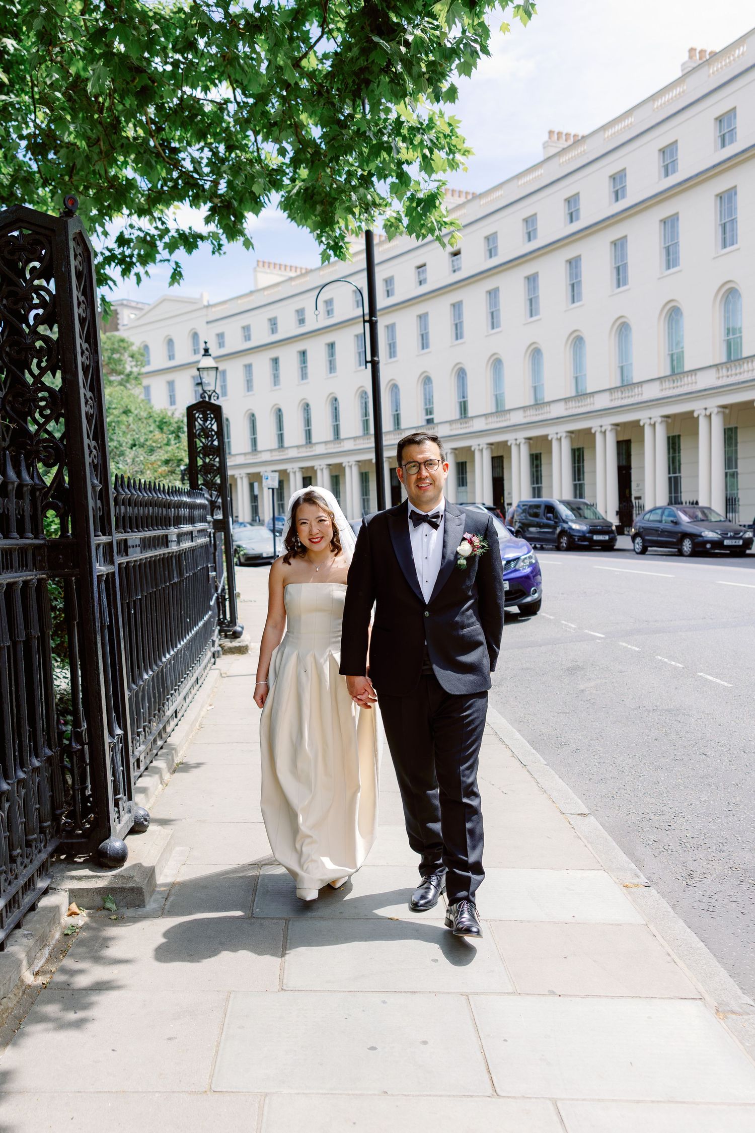 A bride and groom walk hand in hand along a sunlit London street, dressed in elegant wedding attire.