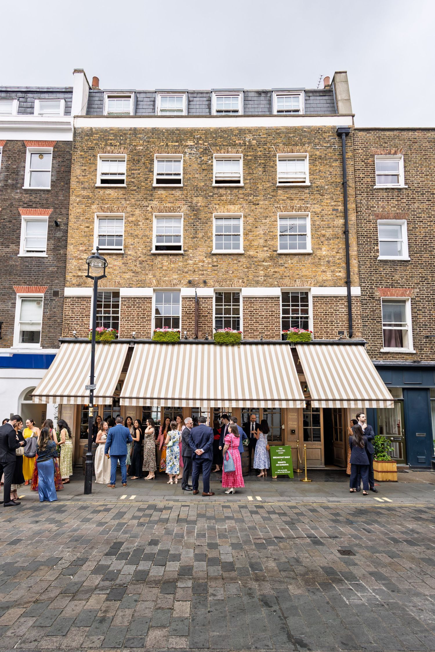 Wedding guests gathering outside a traditional brick building with striped awning on a cobblestone street.