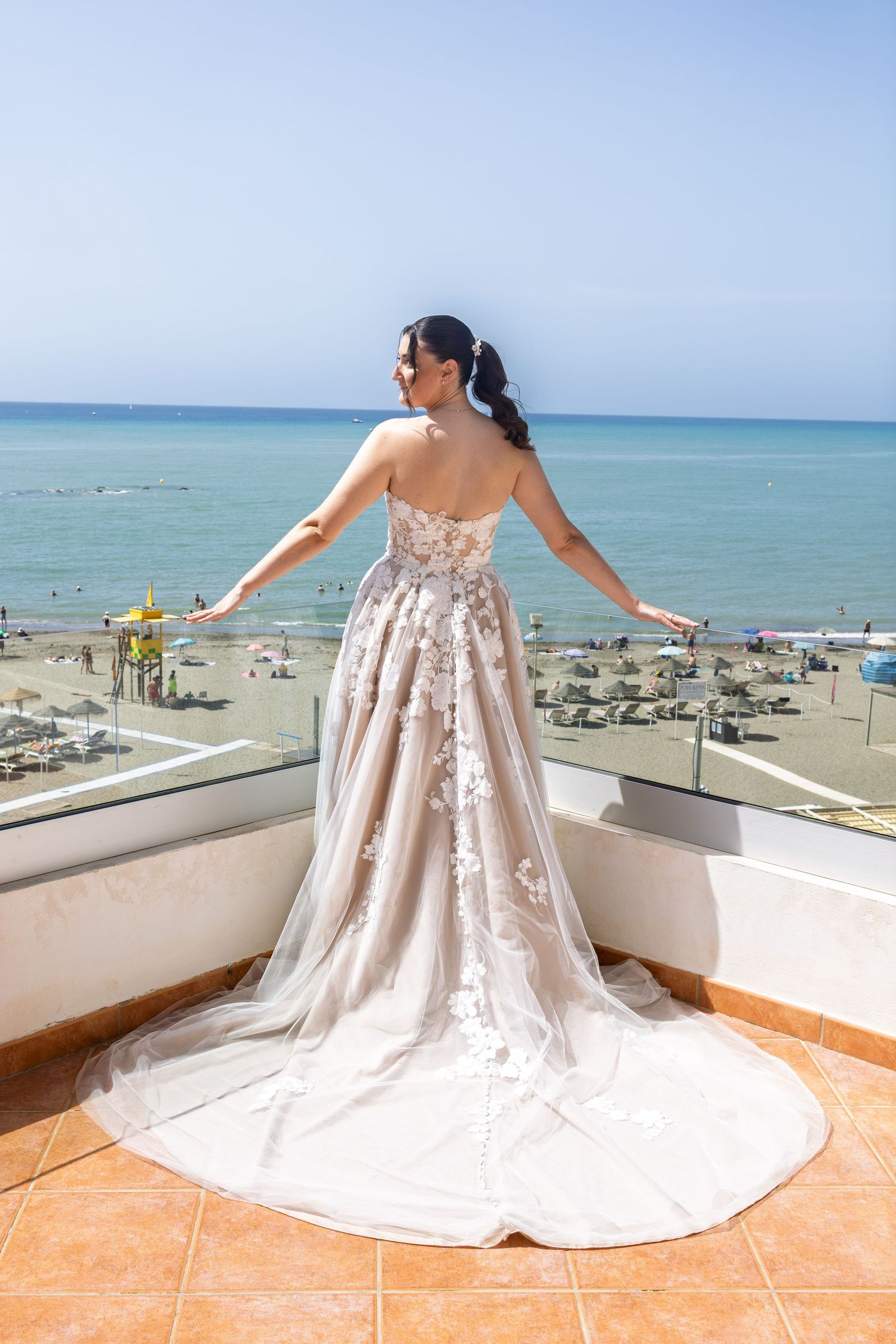 Bride in stunning backless lace wedding gown standing on balcony overlooking a sunny beach and blue sea.