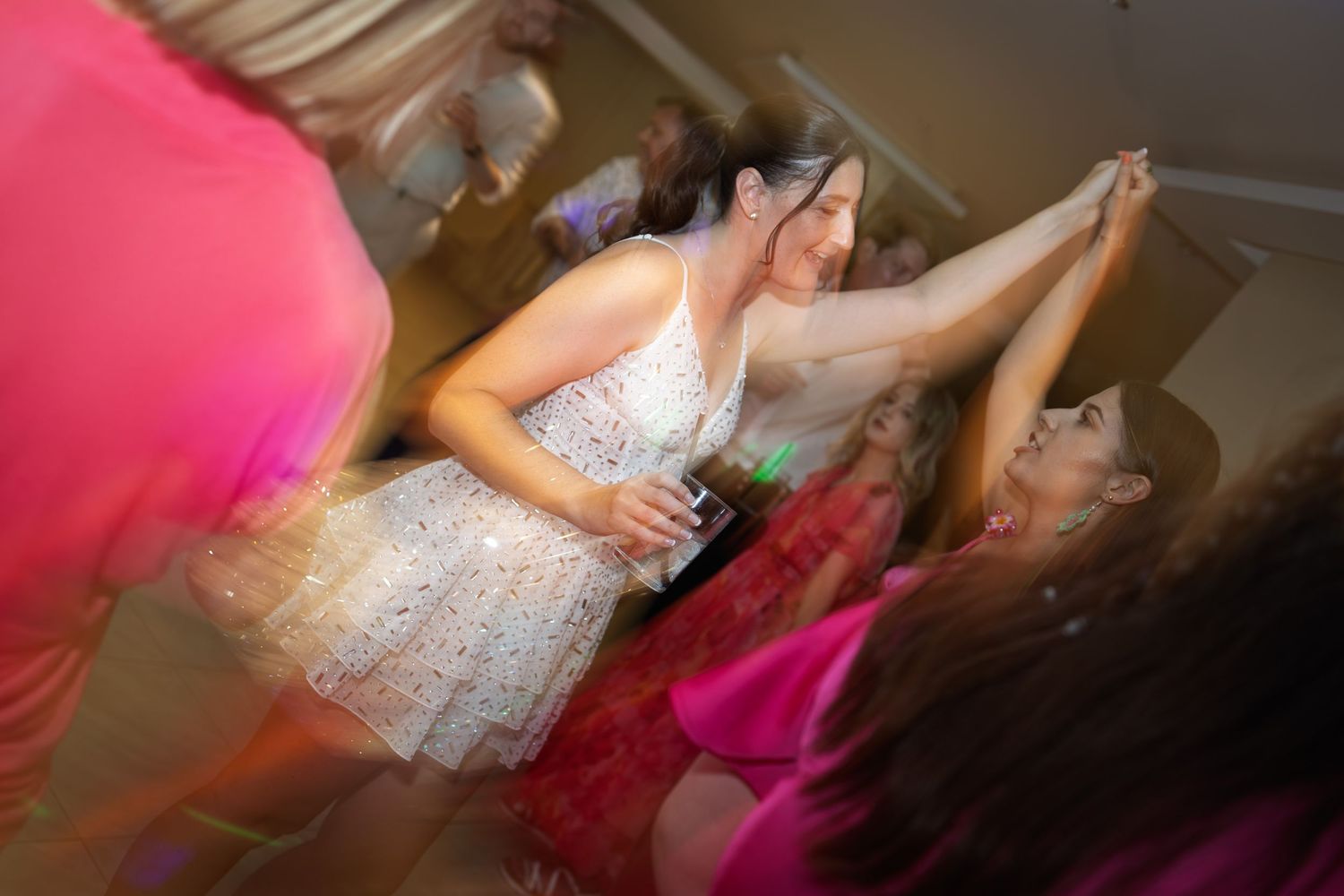 Guests dance energetically at lively wedding reception under colorful lighting in motion-blurred celebration photo.
