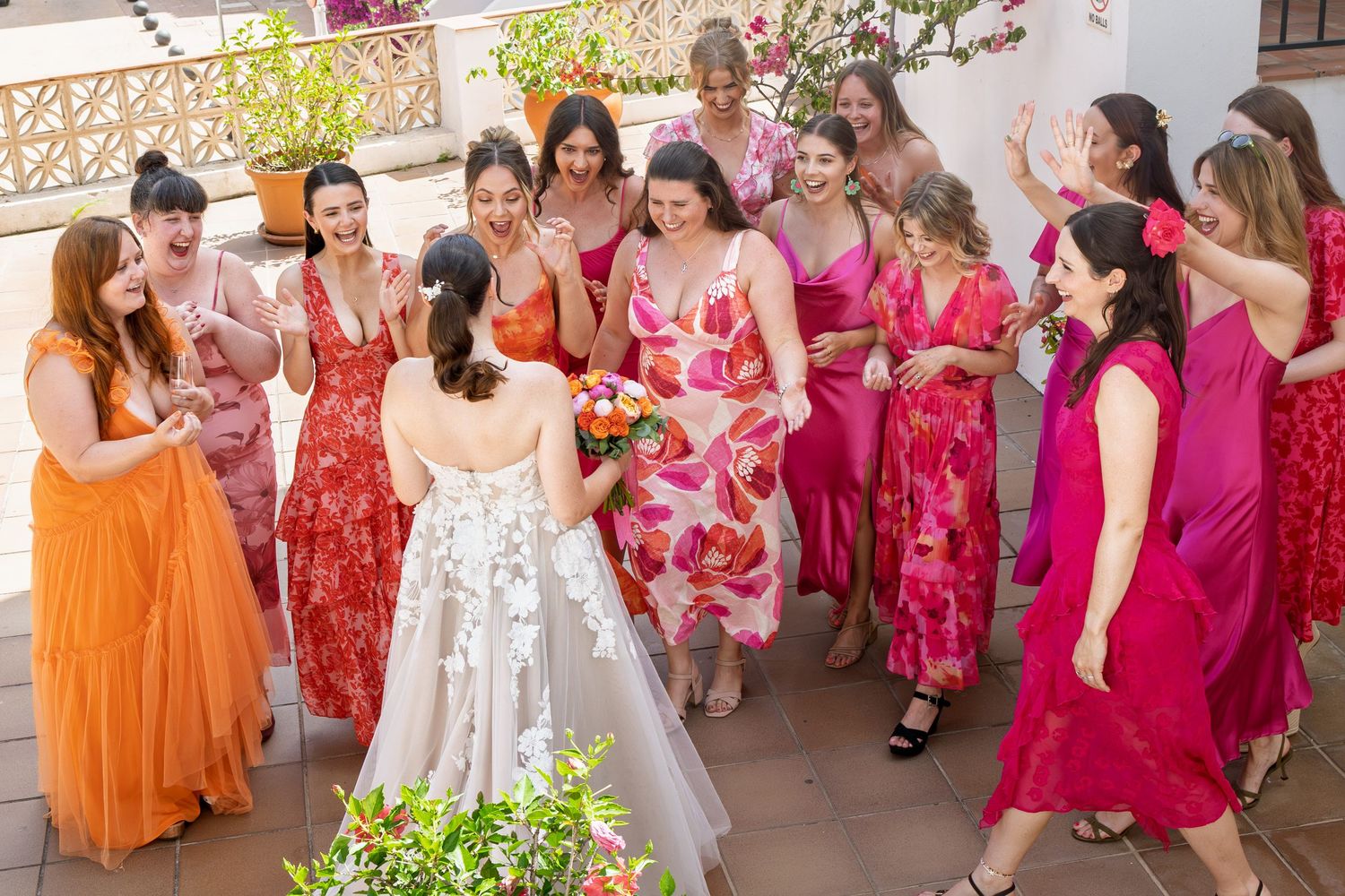 Bride in white gown holds bouquet surrounded by bridesmaids in vibrant red, orange, and pink dresses on a sunny terrace.