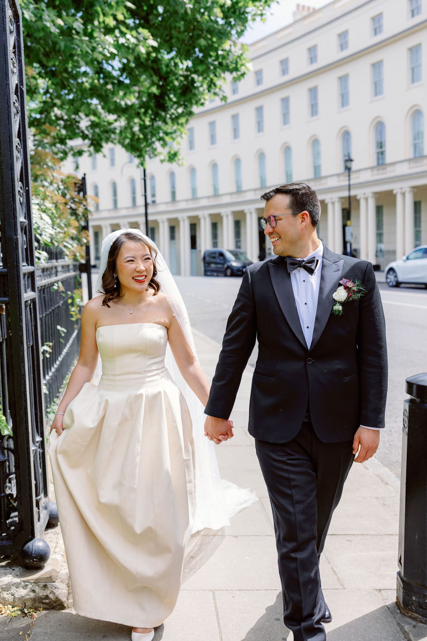 Smiling bride in strapless ivory dress with veil and groom in tuxedo walking together on sunny street.