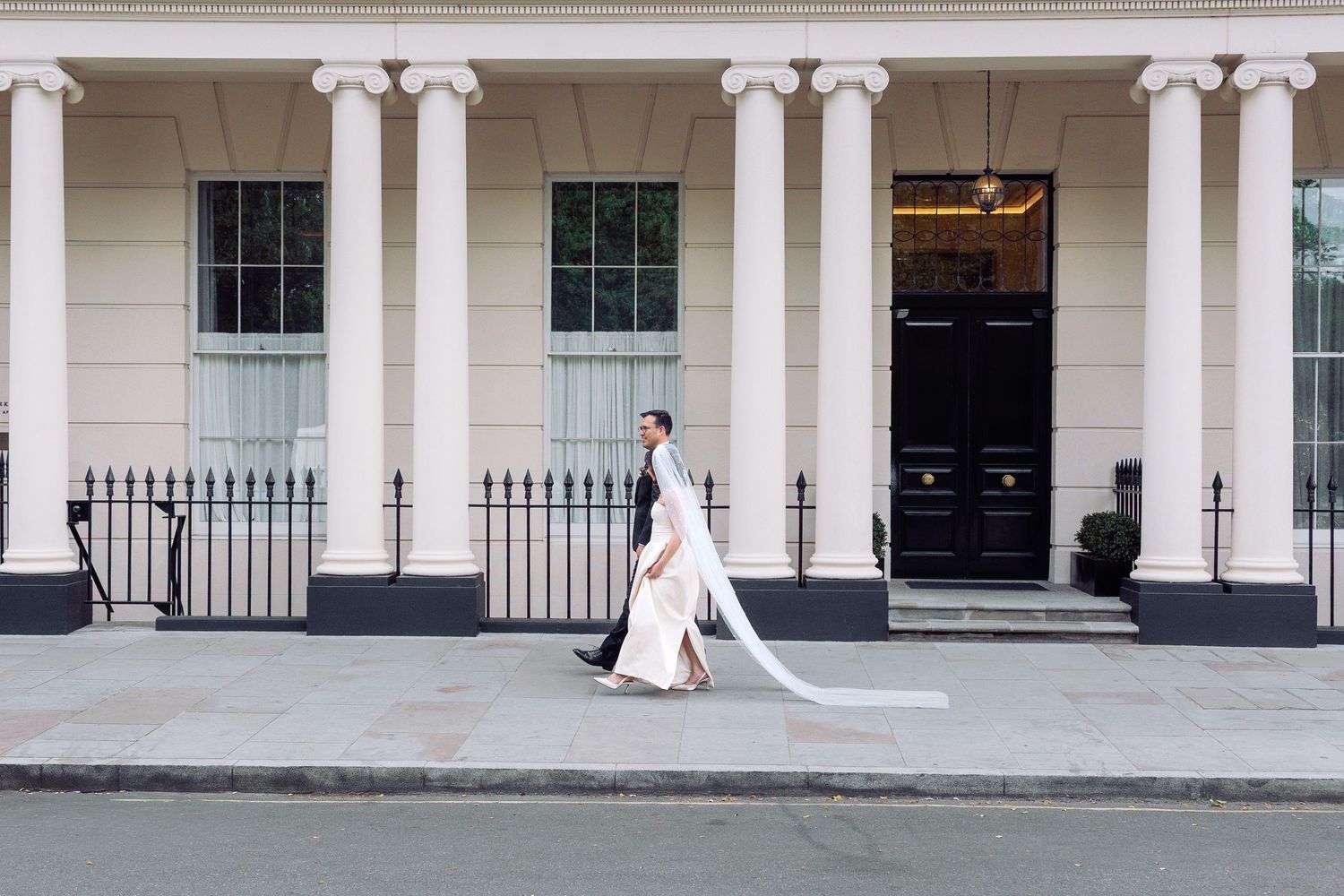 Bride in elegant white wedding dress with long veil walks past classical white columned building.