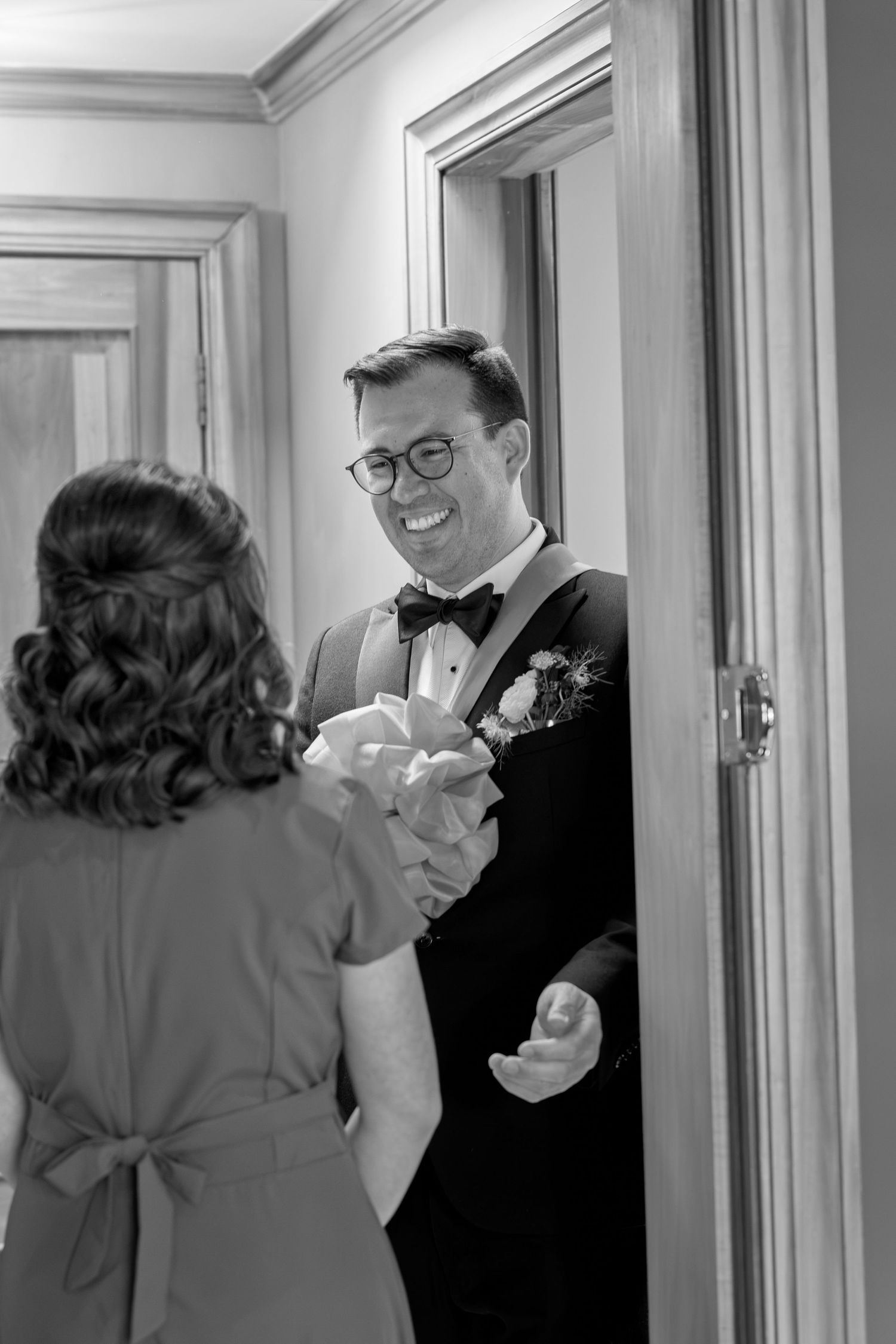 A smiling groom in a tuxedo and bow tie greets a woman at a doorway in black and white.