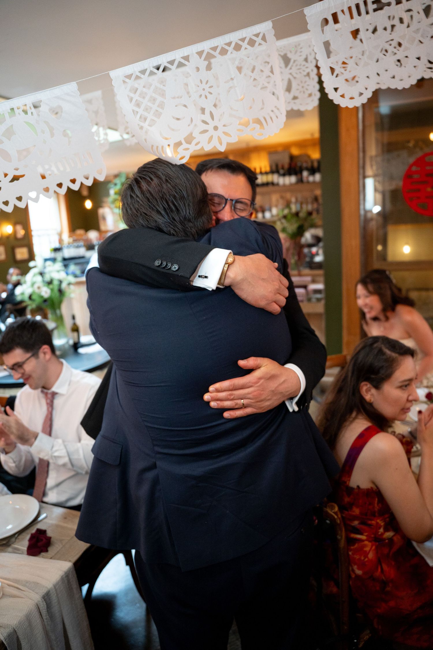 Two men share emotional hug at wedding reception in festive restaurant decorated with paper banners.