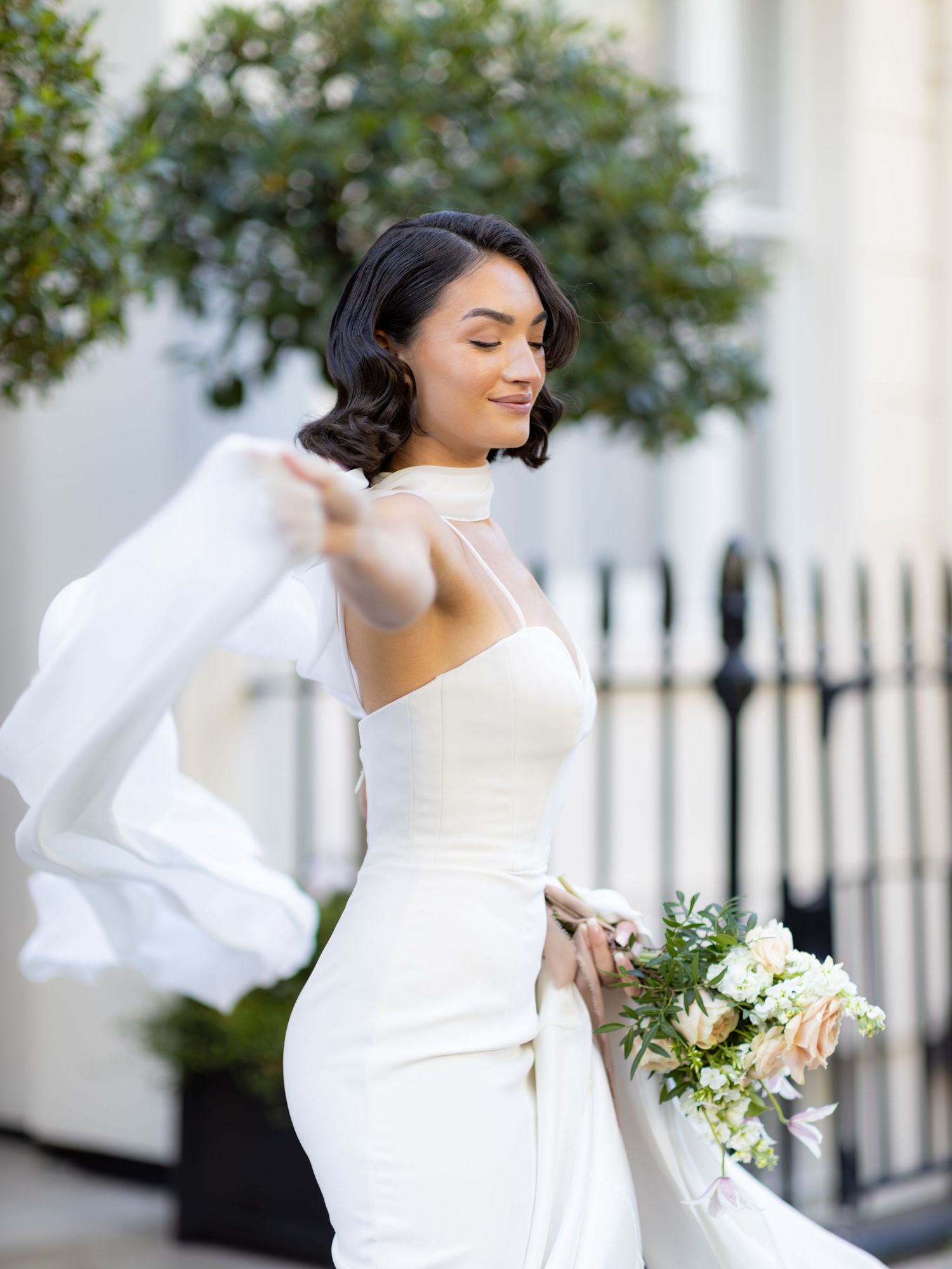 Elegant bride in a white wedding dress twirls joyfully while holding a delicate bouquet outdoors.