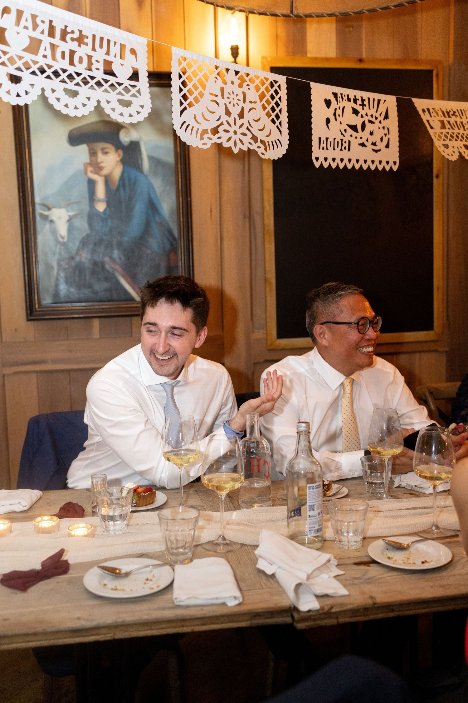 Two men in white shirts laugh together at a festive wedding reception with papel picado decor.