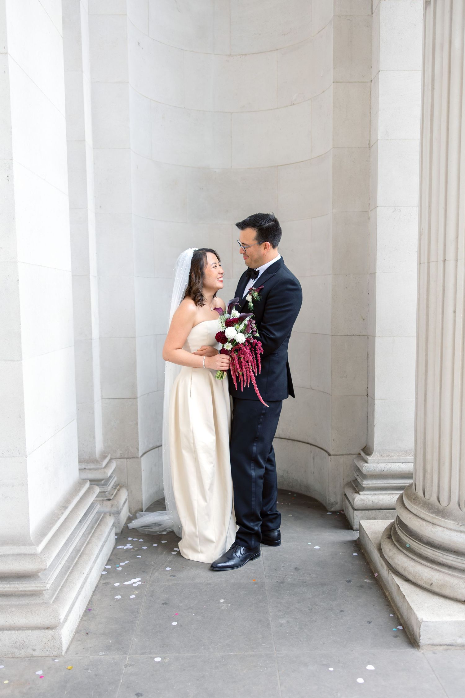Bride and groom share a romantic moment between stone columns holding a burgundy bouquet.