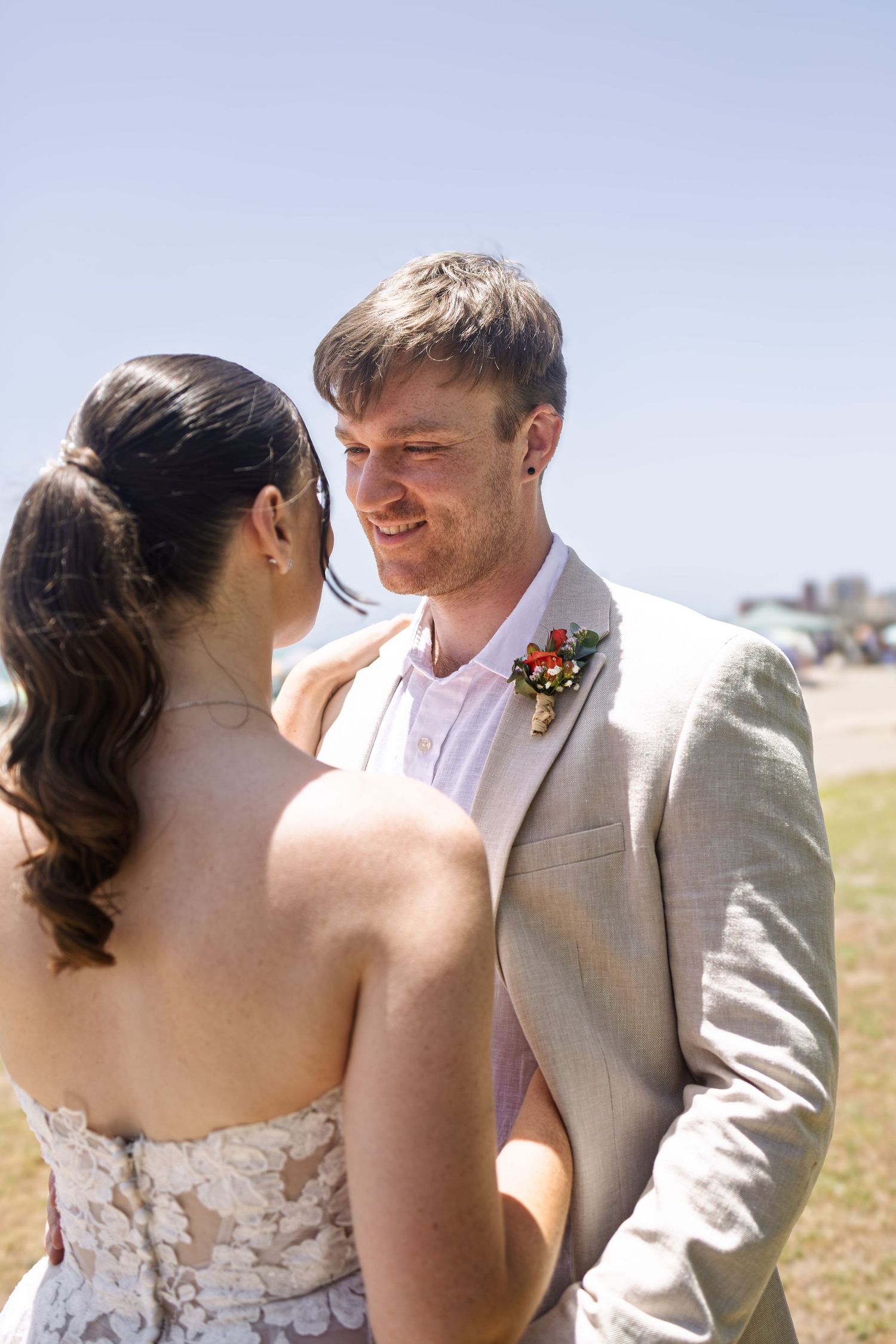 Groom in beige suit with floral boutonniere smiling at bride in lace dress during outdoor beach wedding ceremony.