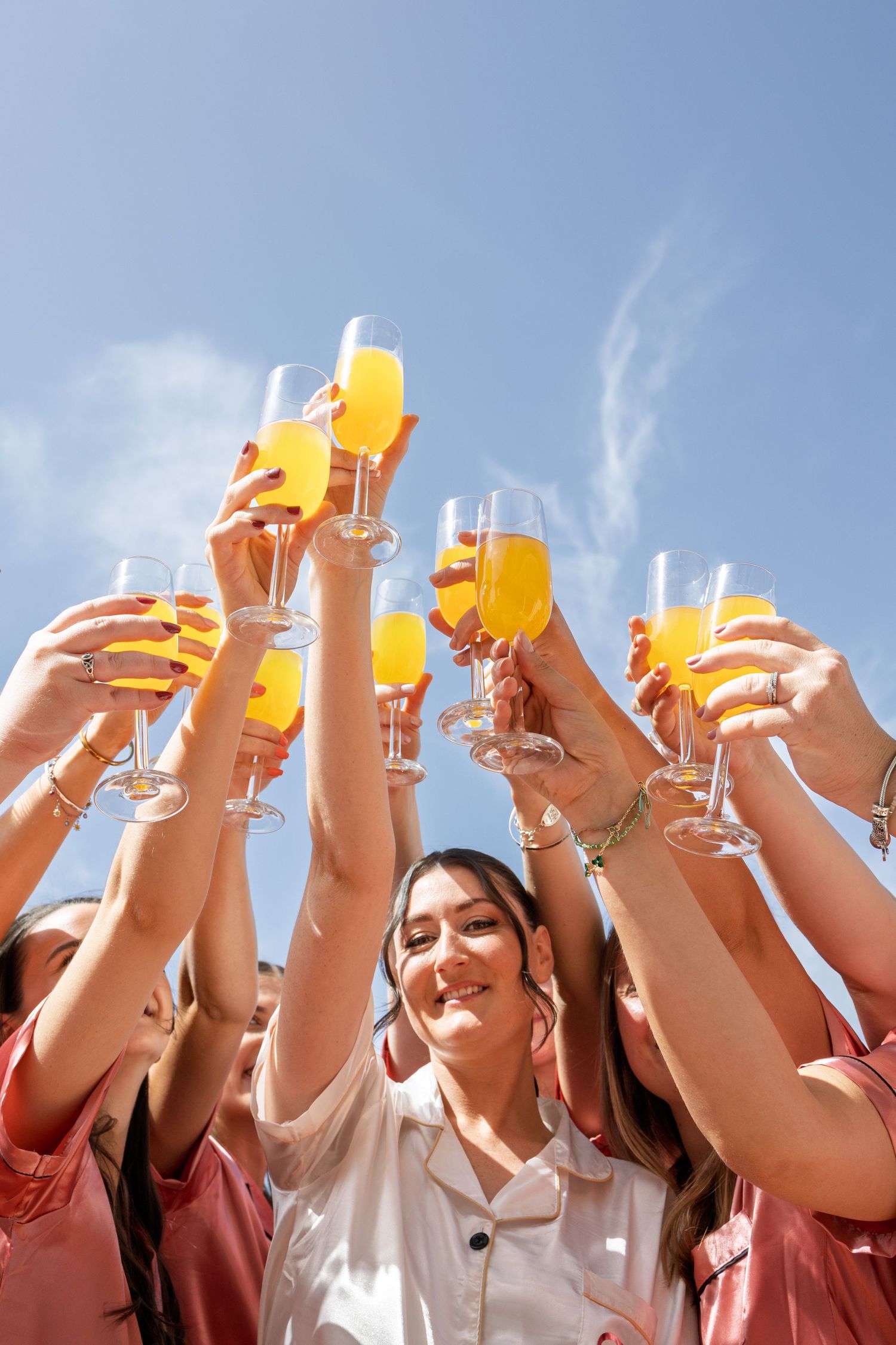 Group of women raising champagne flutes with orange juice in a celebratory toast against a blue sky.