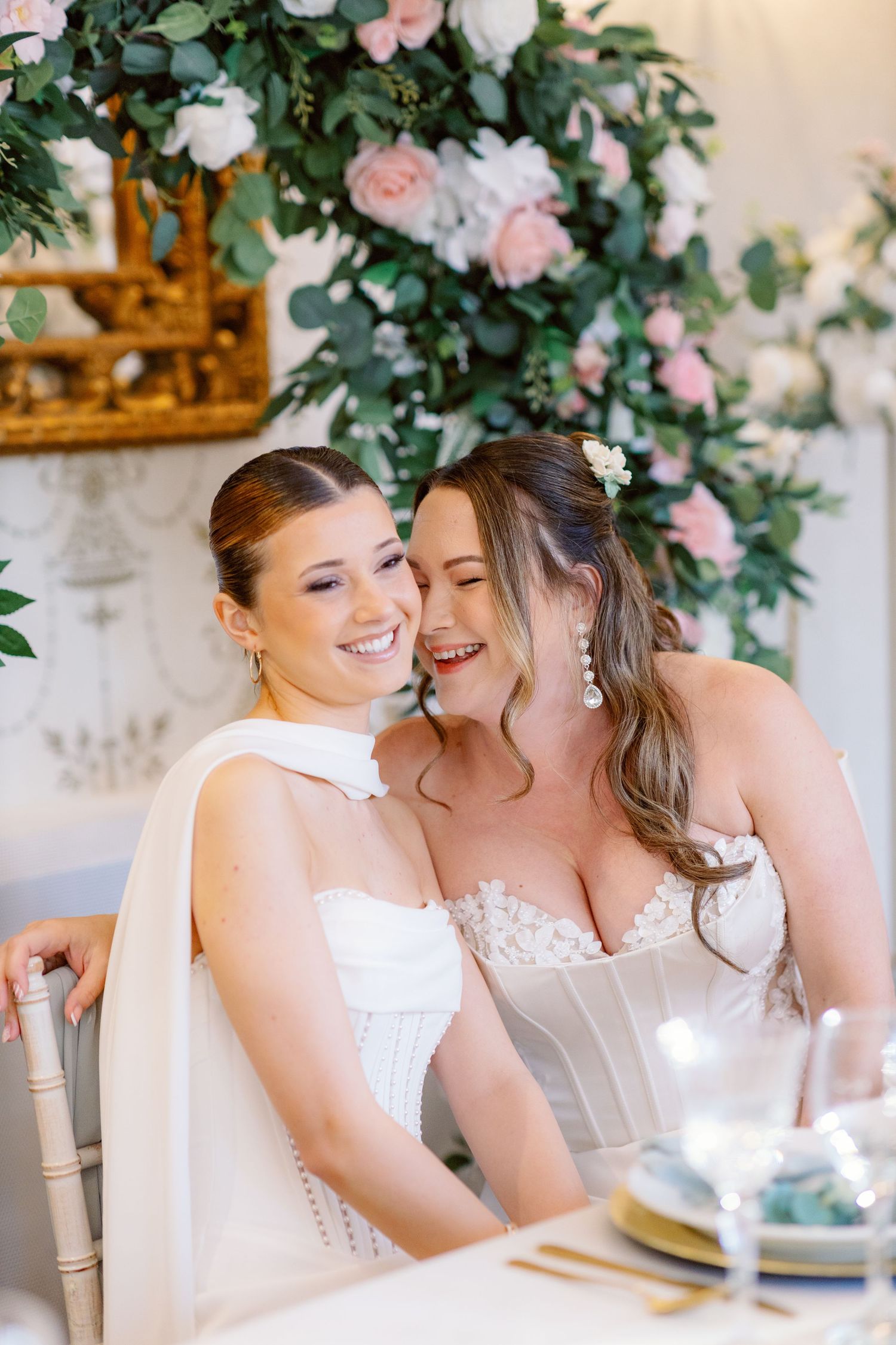 Two people in white dresses share a joyful moment at a wedding reception decorated with pink and white floral arrangements.