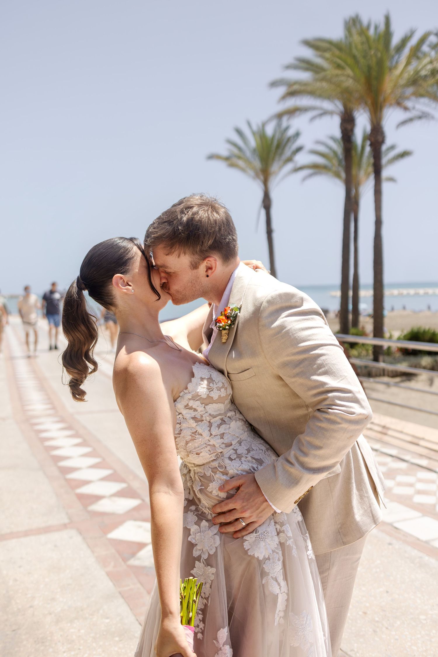 Newlywed couple sharing a kiss on a sunny promenade lined with tall palm trees near the beach.