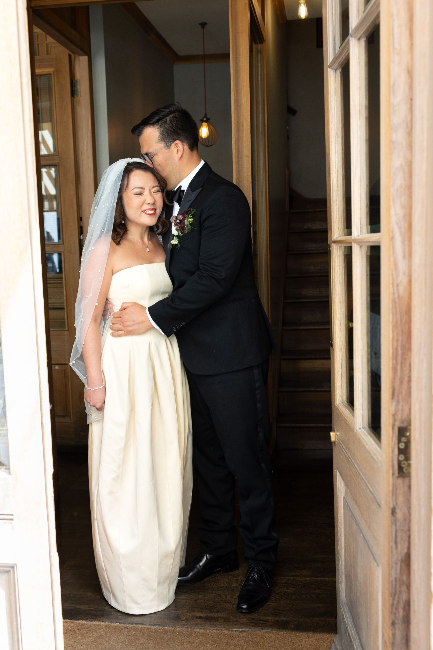 Asian bride and groom sharing intimate moment in doorway, bride in strapless white gown with veil, groom in black tuxedo.