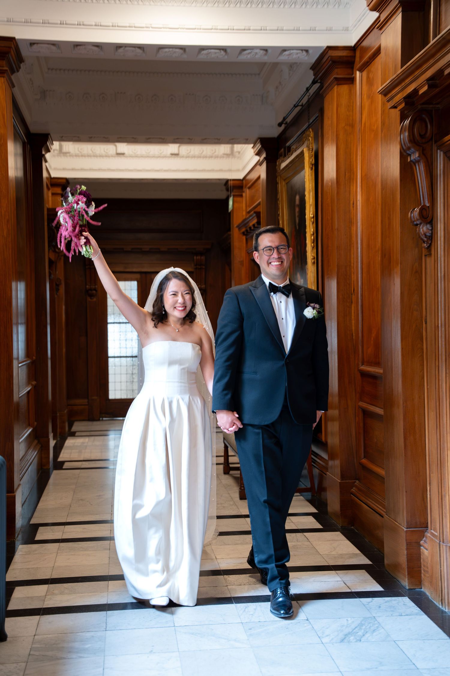 Joyful newlyweds walk hand-in-hand through grand wood-paneled hall, bride raising bouquet in celebration.