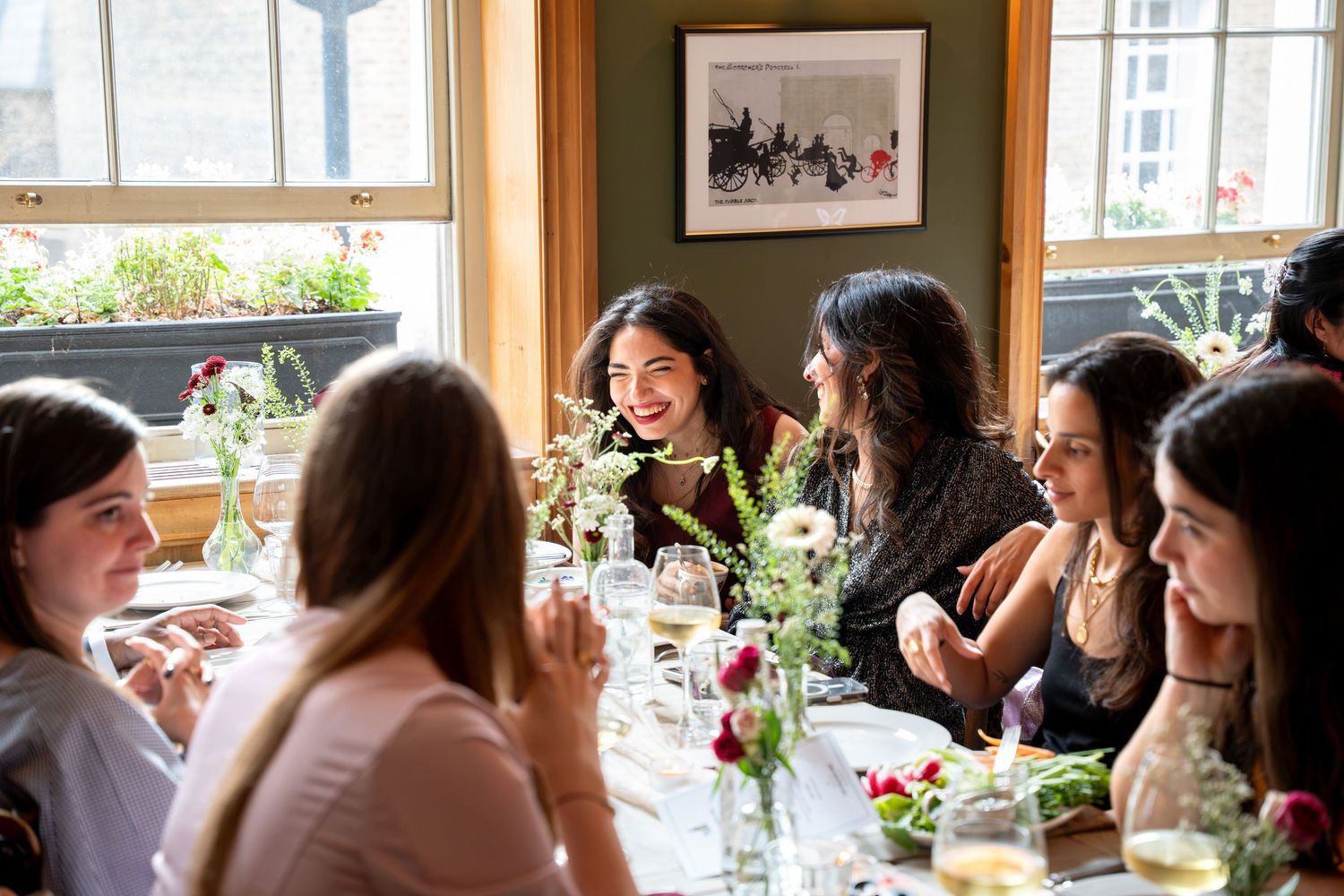 Group of women laughing and celebrating together at a sunlit wedding reception dining table with florals.