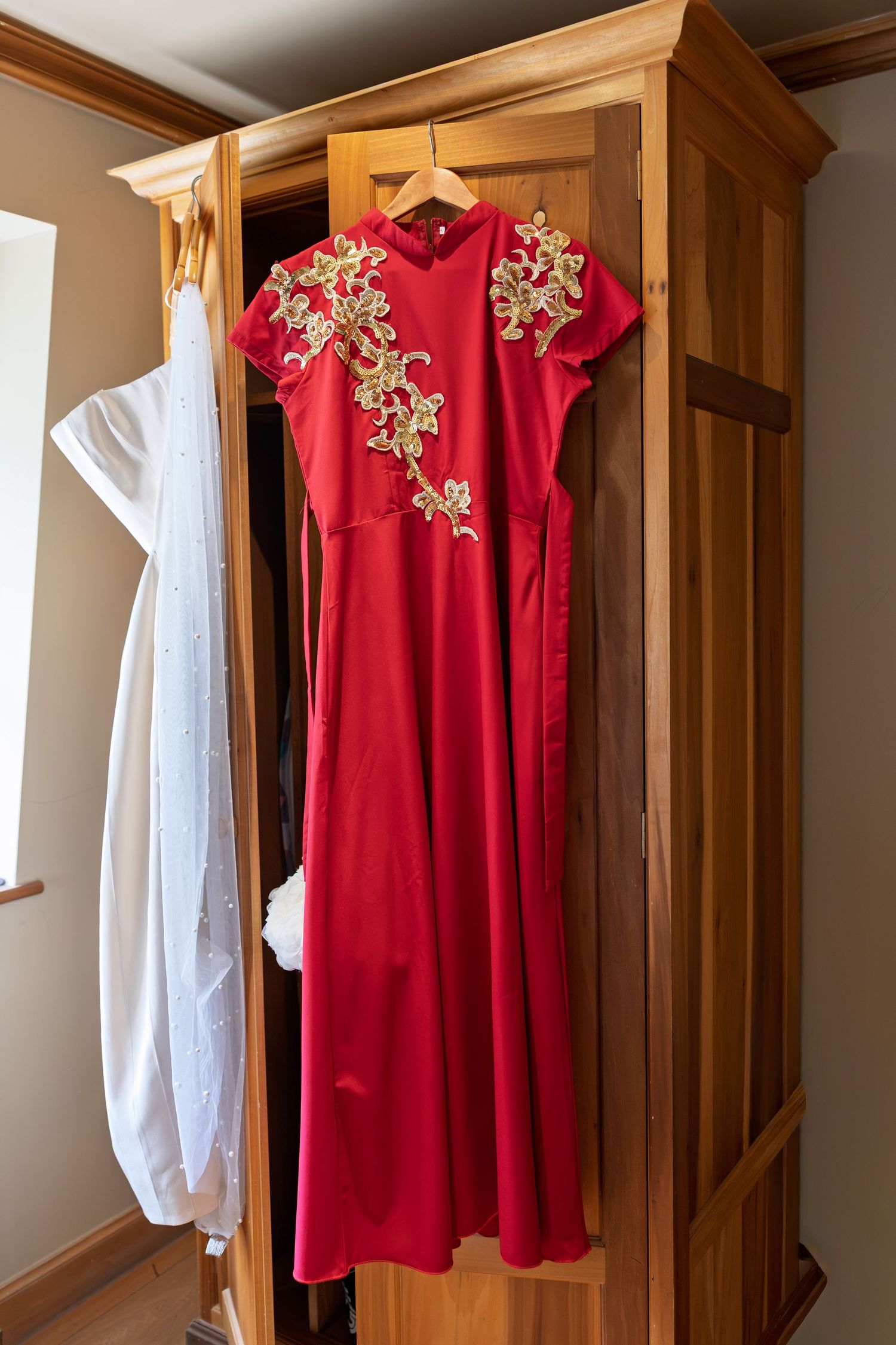 Red Chinese qipao dress with gold floral embroidery hanging on wooden wardrobe door.