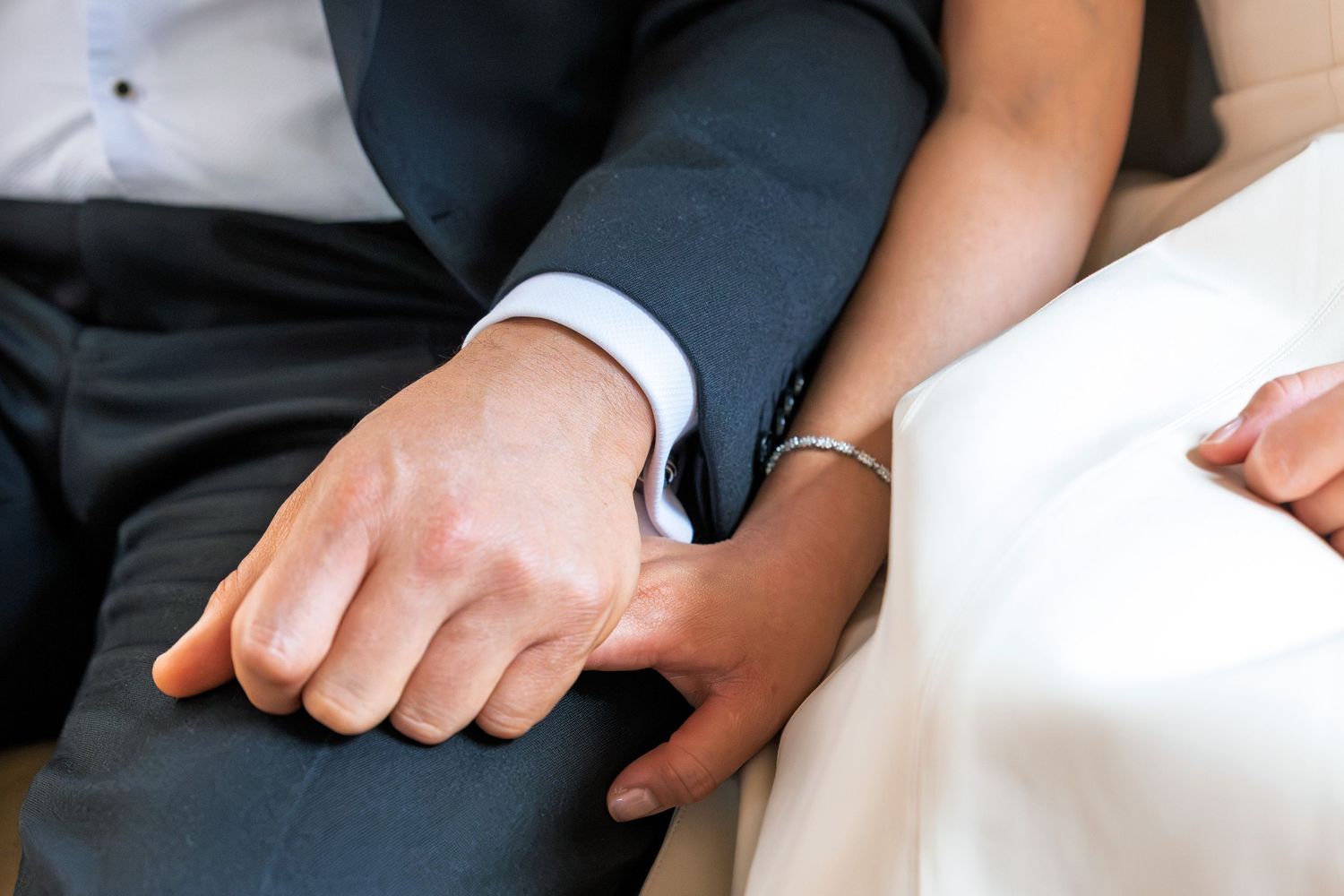Close-up of bride and groom holding hands, bride wearing bracelet, groom in navy suit, at wedding ceremony.