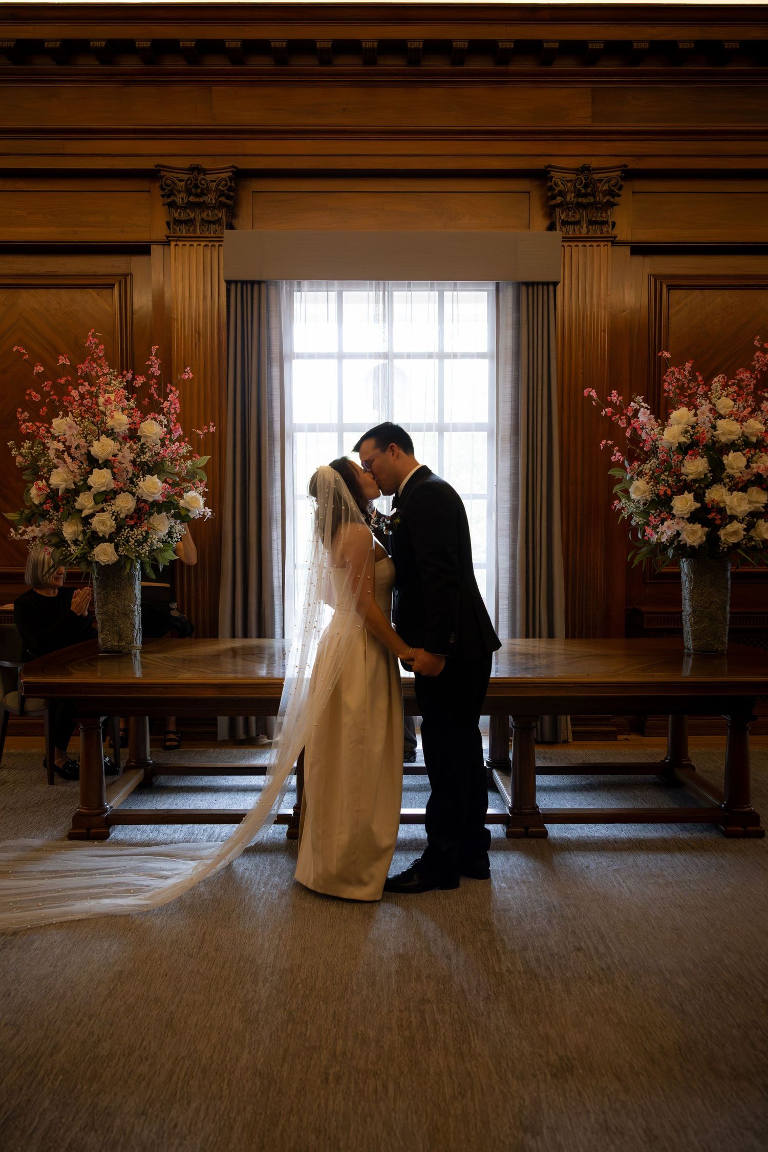 Bride and groom share first kiss at elegant wedding ceremony with floral arrangements and wood paneling.