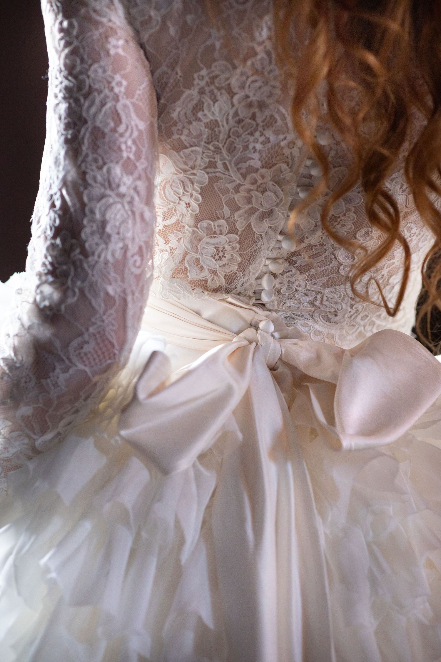 Close-up detail of white wedding dress showing intricate lace pattern and satin bow.