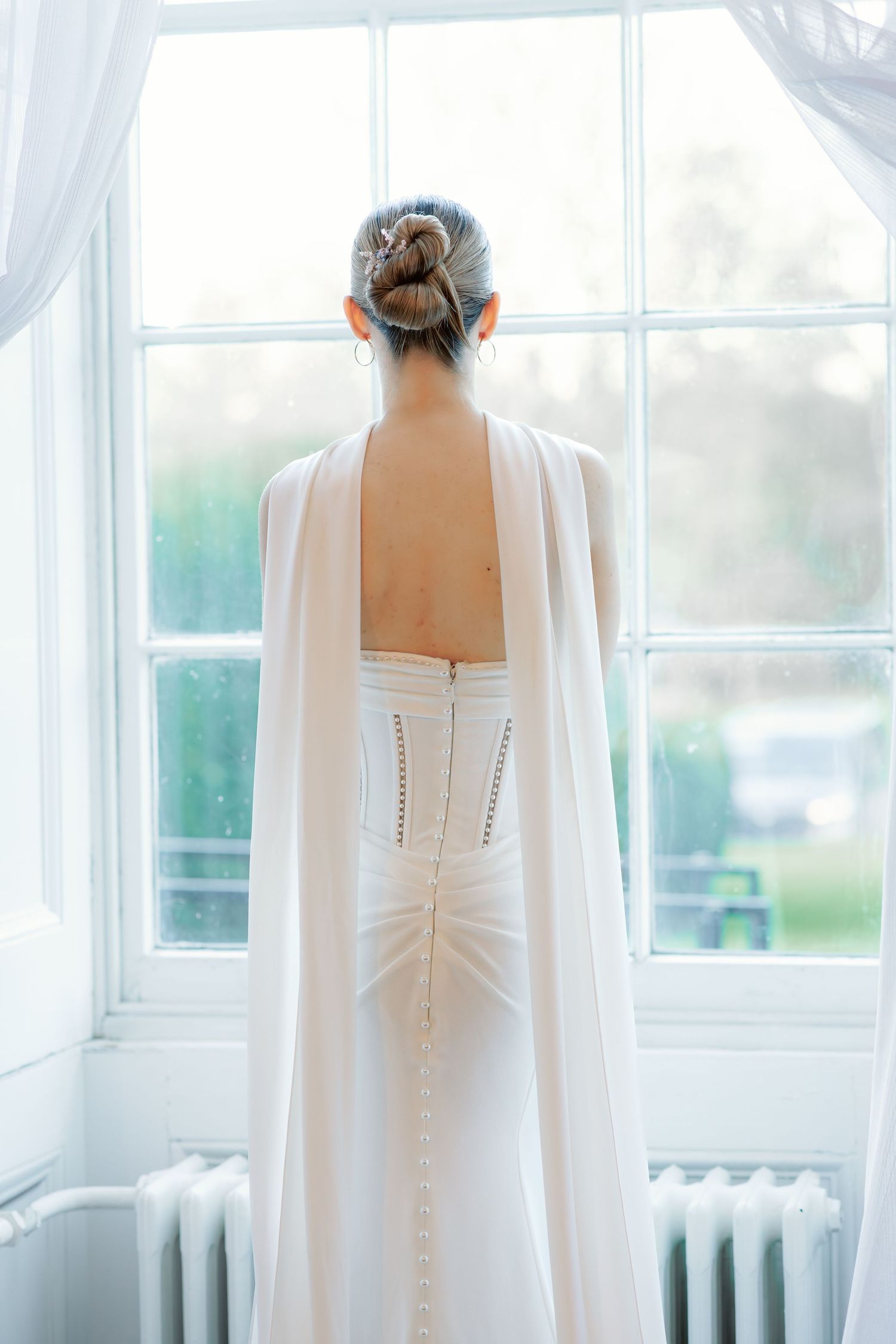 Wedding dress details showing buttons and lace on back with white cape against bright windows.
