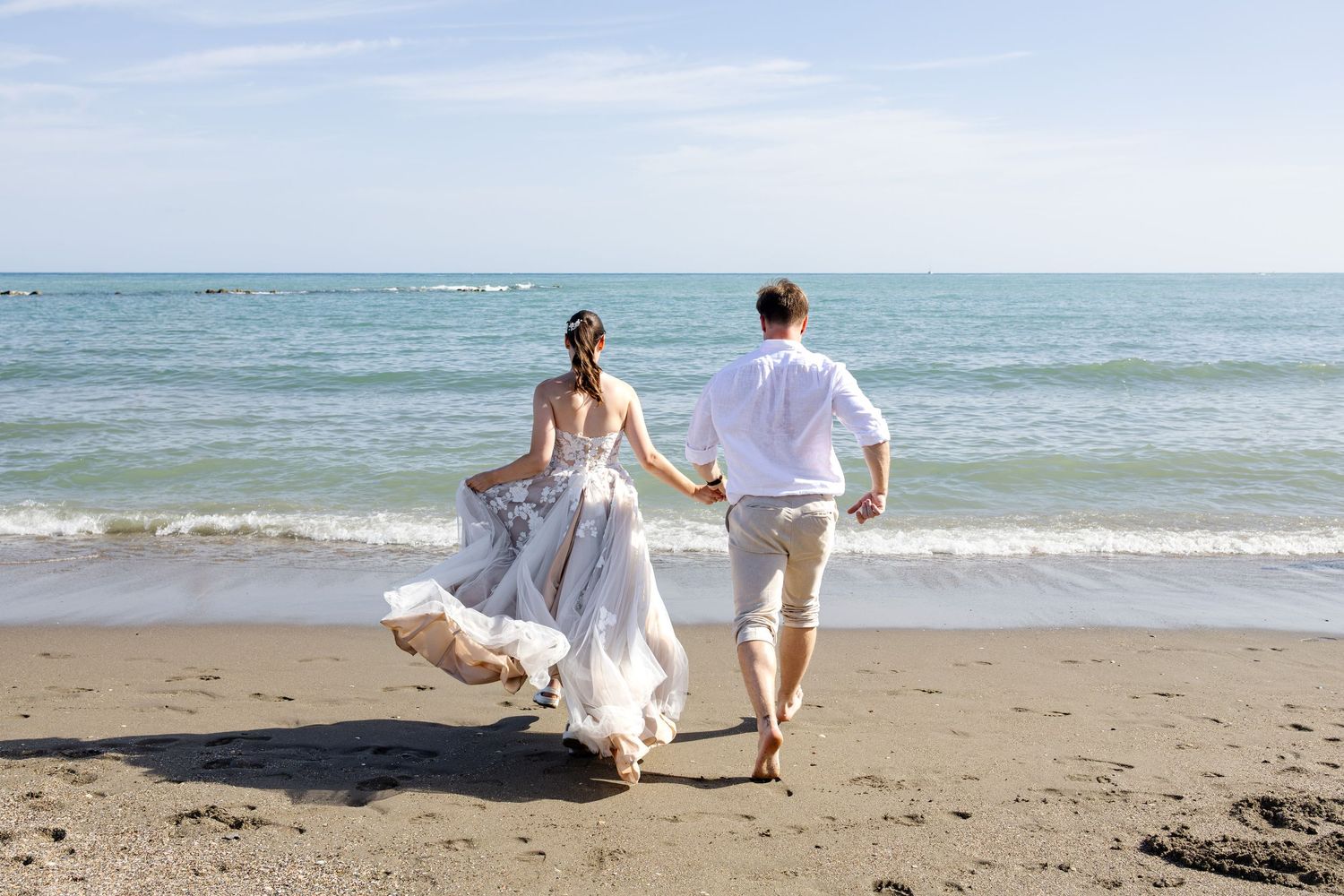 Bride and groom running hand in hand toward ocean waves on a beautiful sunny beach.