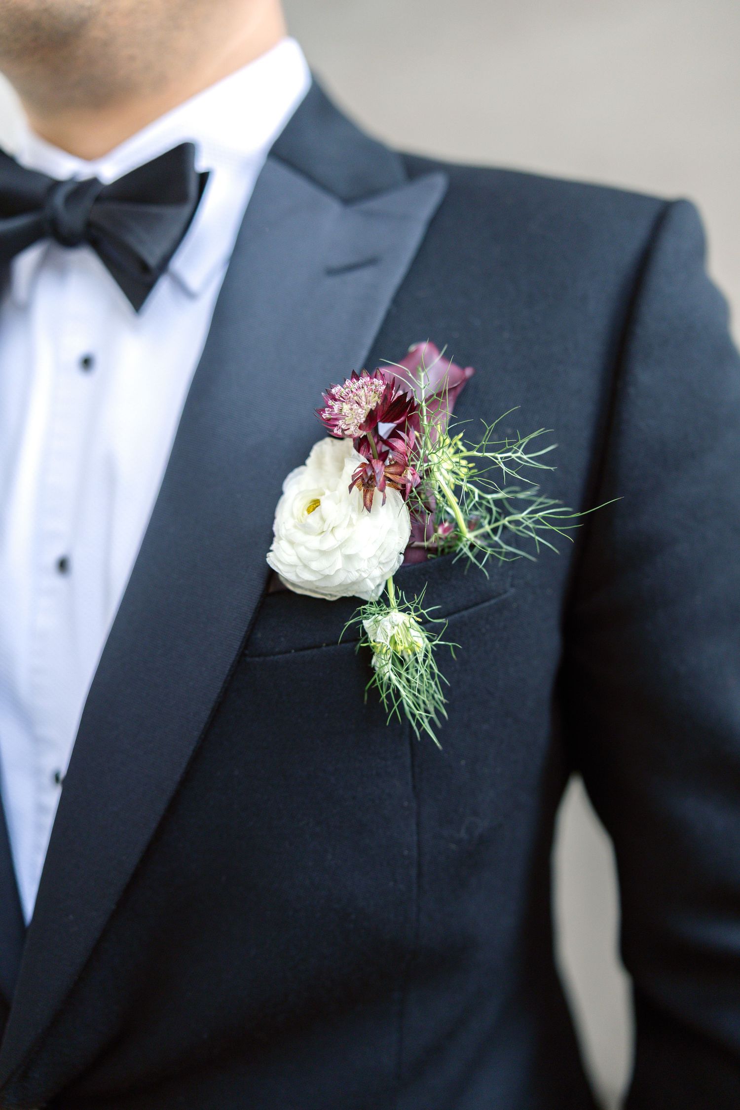 Close-up of a groom's boutonniere featuring white ranunculus and burgundy flowers on a black tuxedo lapel.