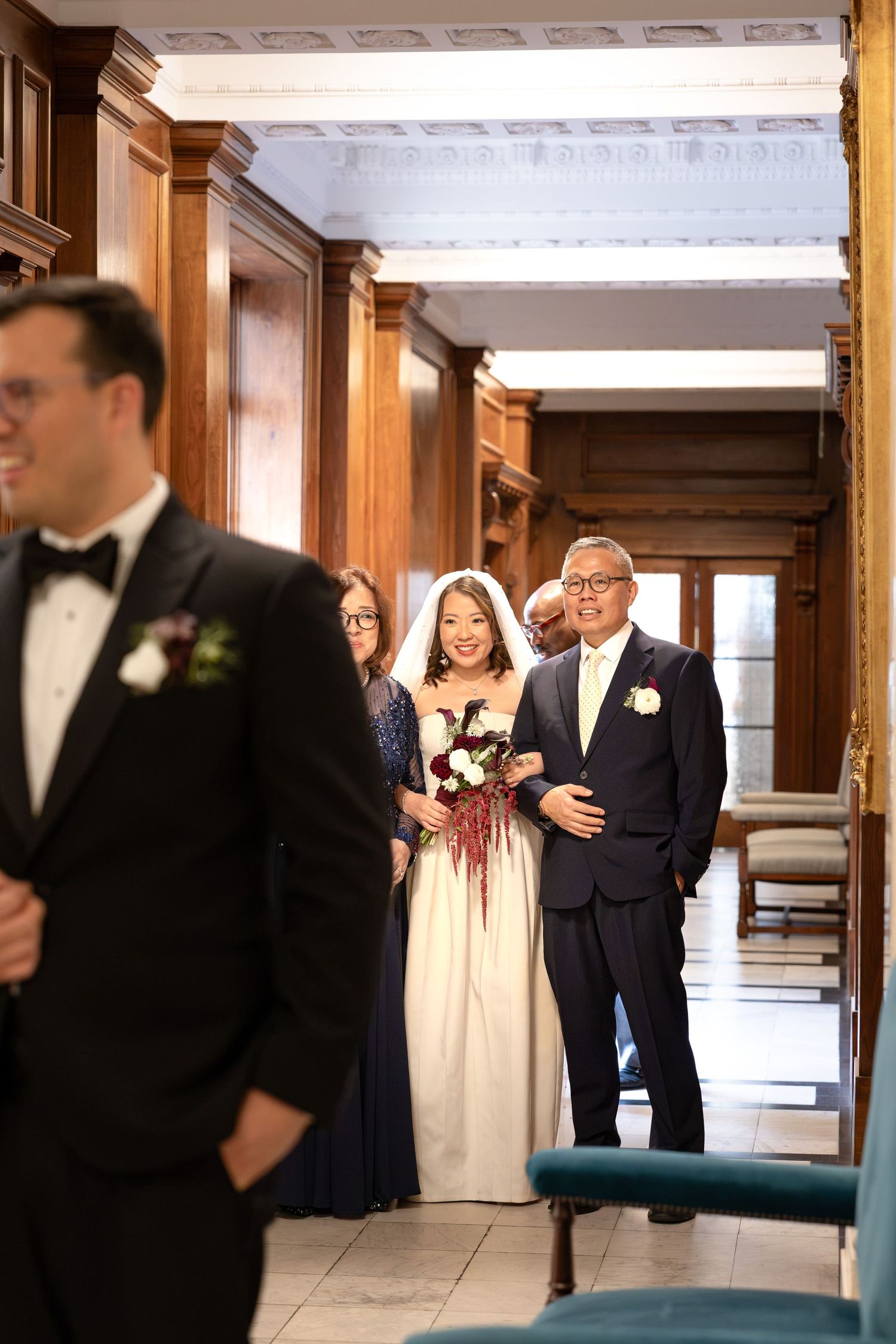 Bride walking down the aisle with her father in an elegant wood-paneled venue holding a red bouquet.