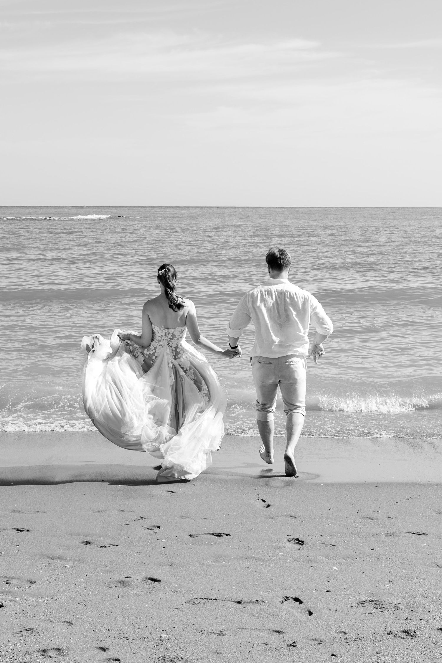 Black and white photo of bride and groom running hand in hand toward the ocean on a sandy beach.