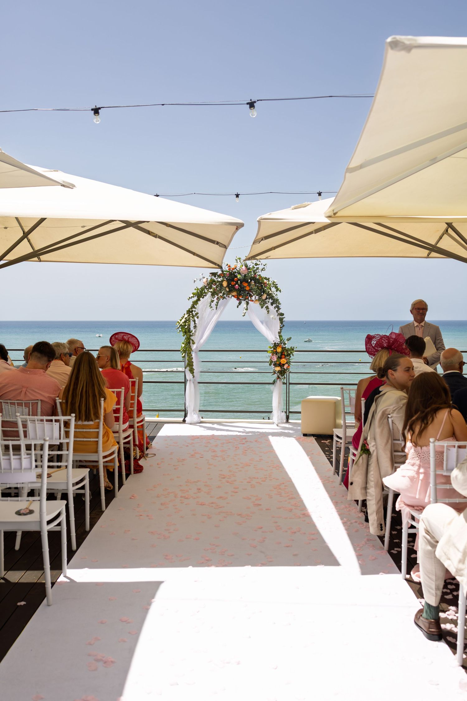 Elegant beachside wedding ceremony setup with white aisle, floral arch, and guests seated under large umbrellas.