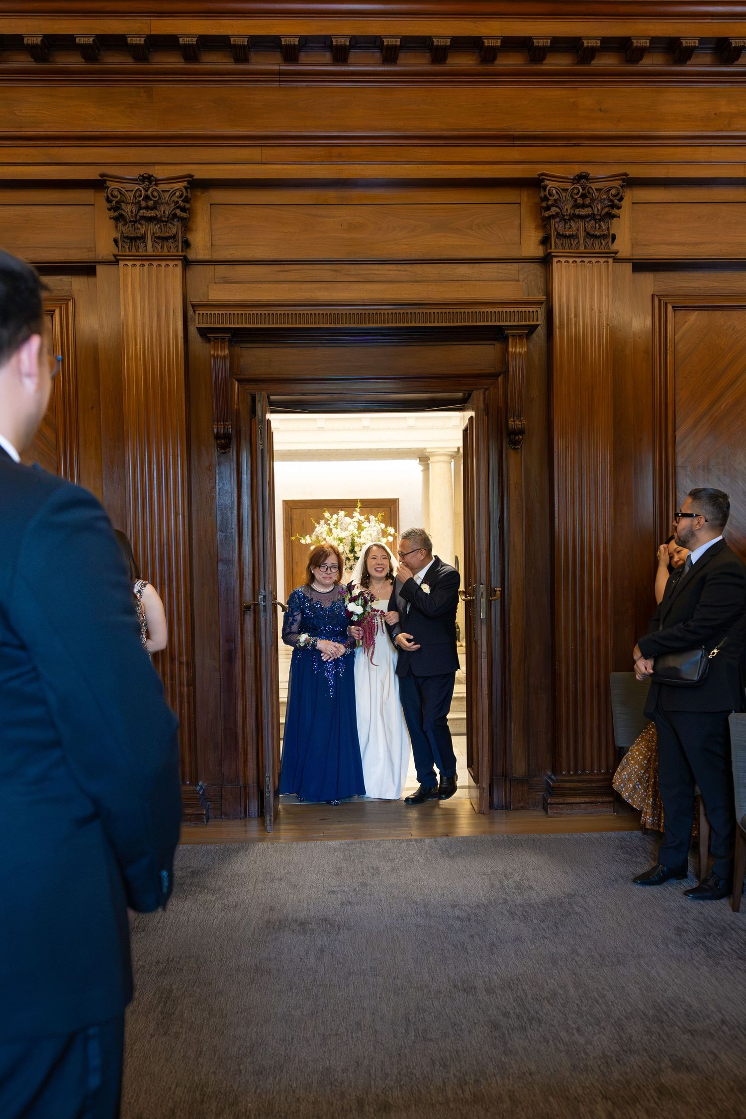 Bride escorted by parents through ornate wooden doors into a grand wedding ceremony venue.