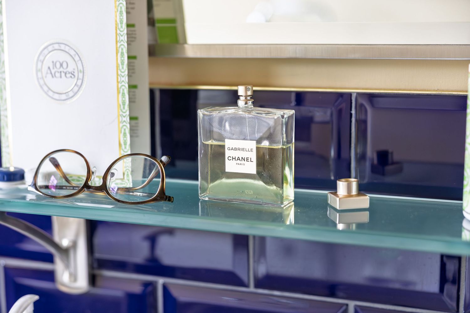 Chanel Gabrielle perfume bottle displayed on a glass shelf beside eyeglasses and a small fragrance cap.