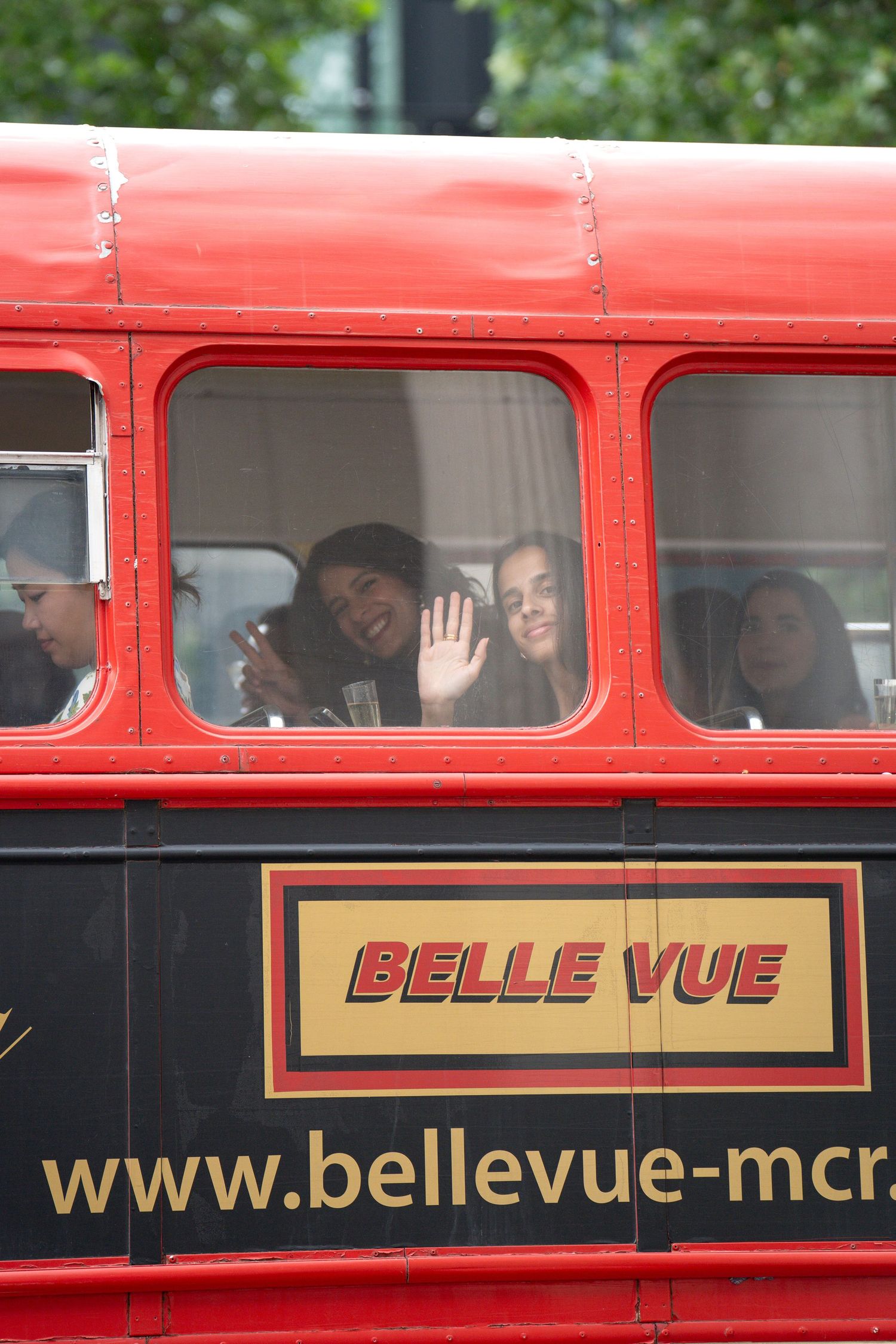 Wedding guests wave and smile from the windows of a red Belle Vue double-decker bus.