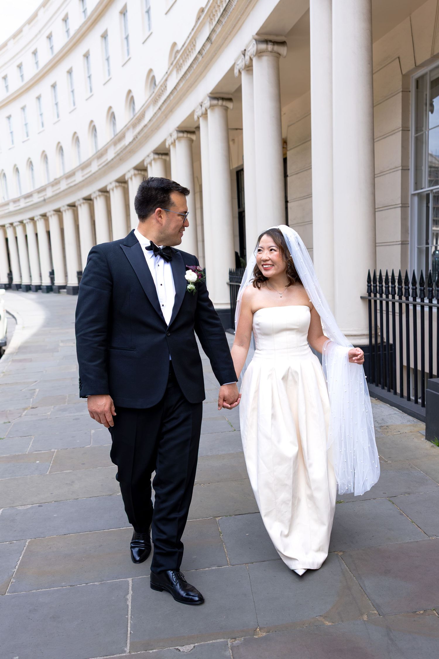 Elegant bride and groom walk hand in hand beneath curved white colonnaded building in London.