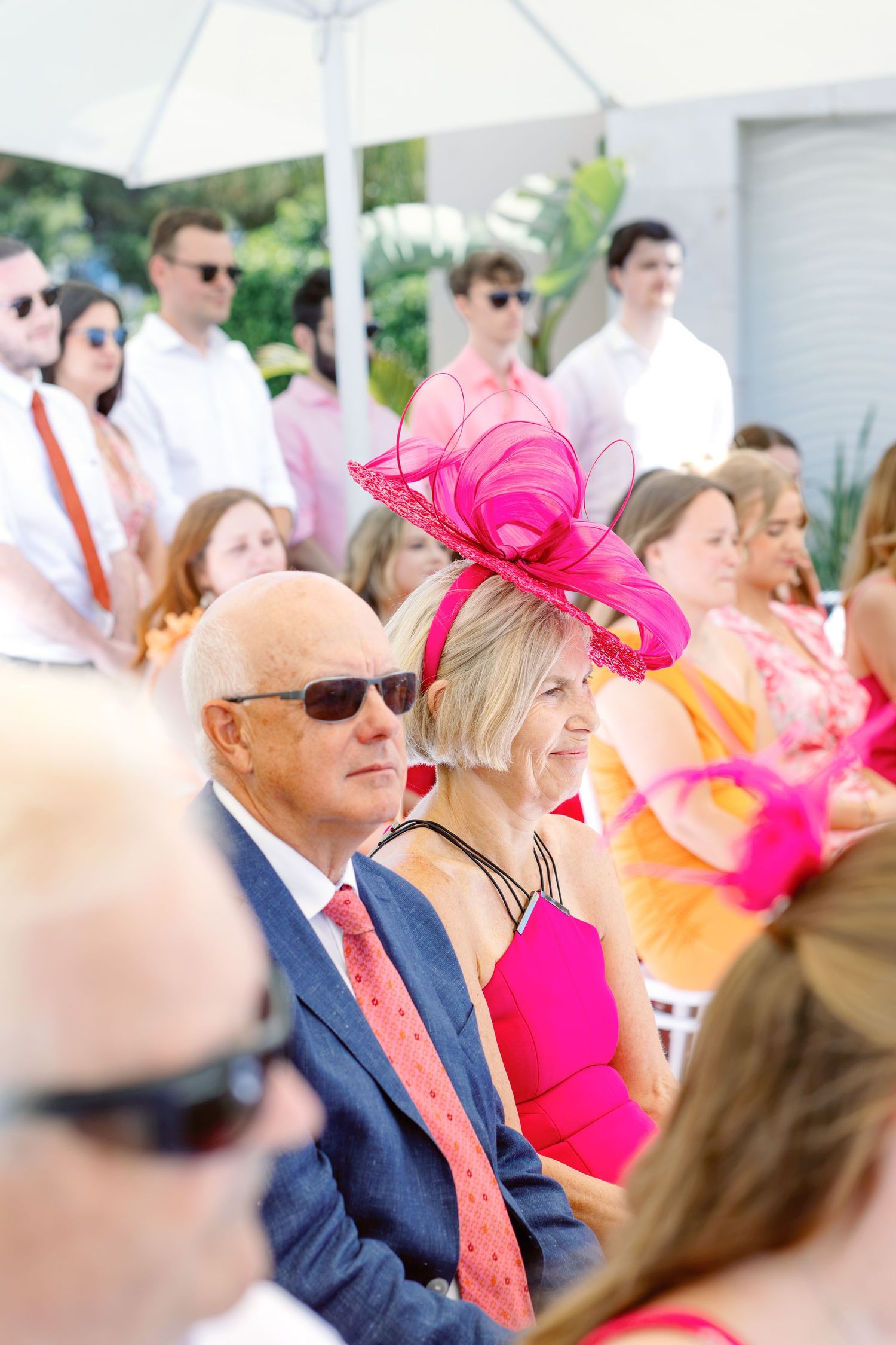 Elegantly dressed guests wearing vibrant pink fascinators and hats at an outdoor garden party event.