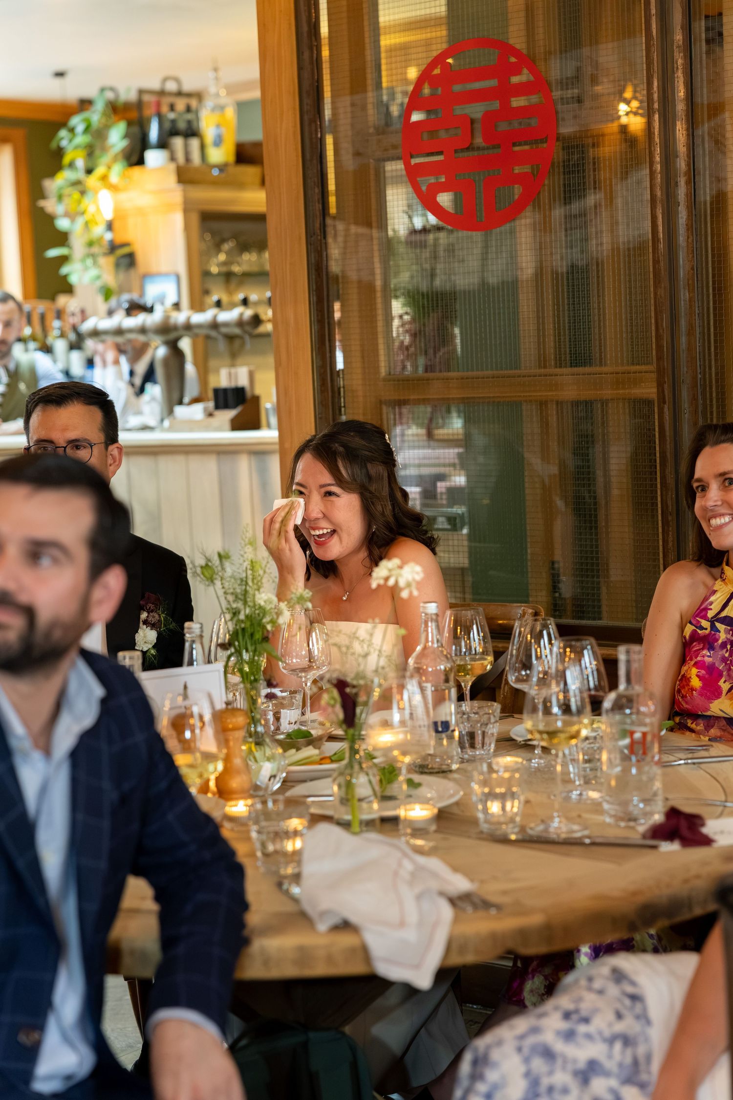 Bride laughs joyfully at wedding reception dinner table decorated with Chinese double happiness symbol.