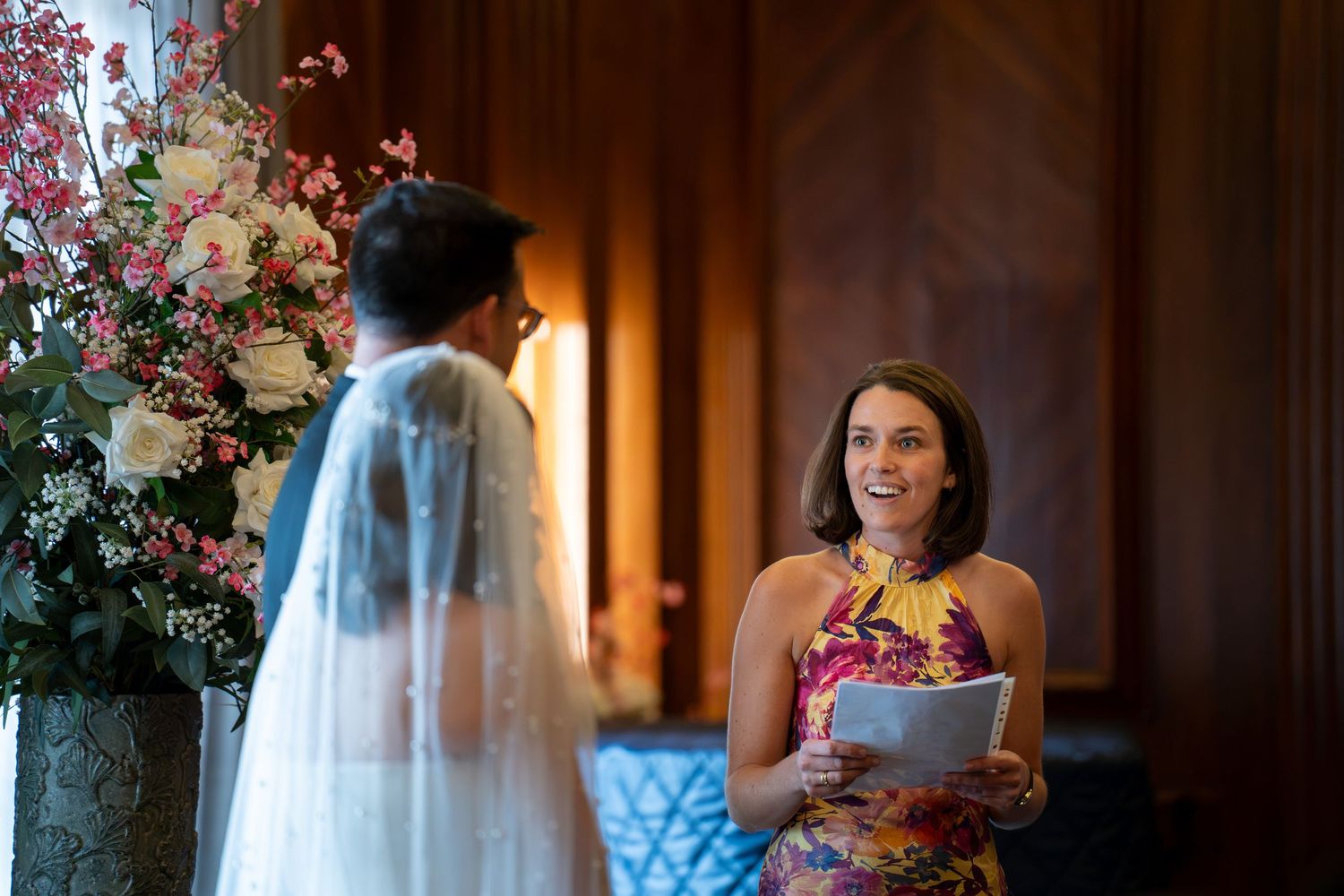 Wedding officiant smiling warmly at bride during intimate ceremony beside floral arrangements.