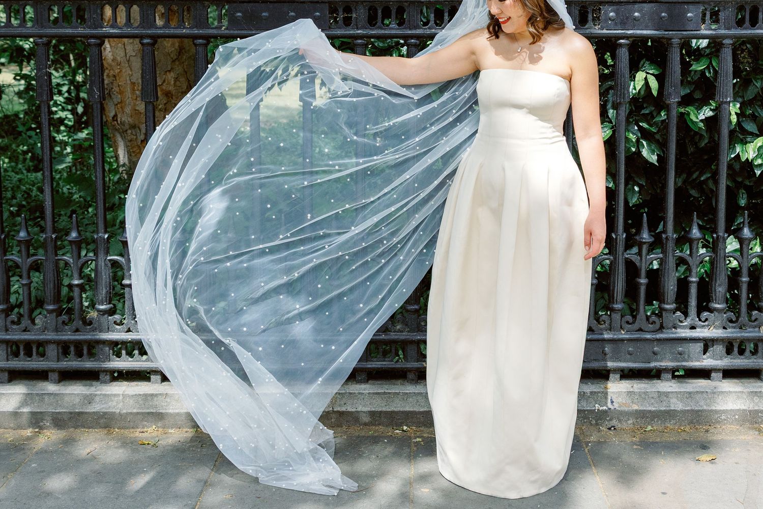Bride in white strapless gown with flowing veil outdoors near iron fence.