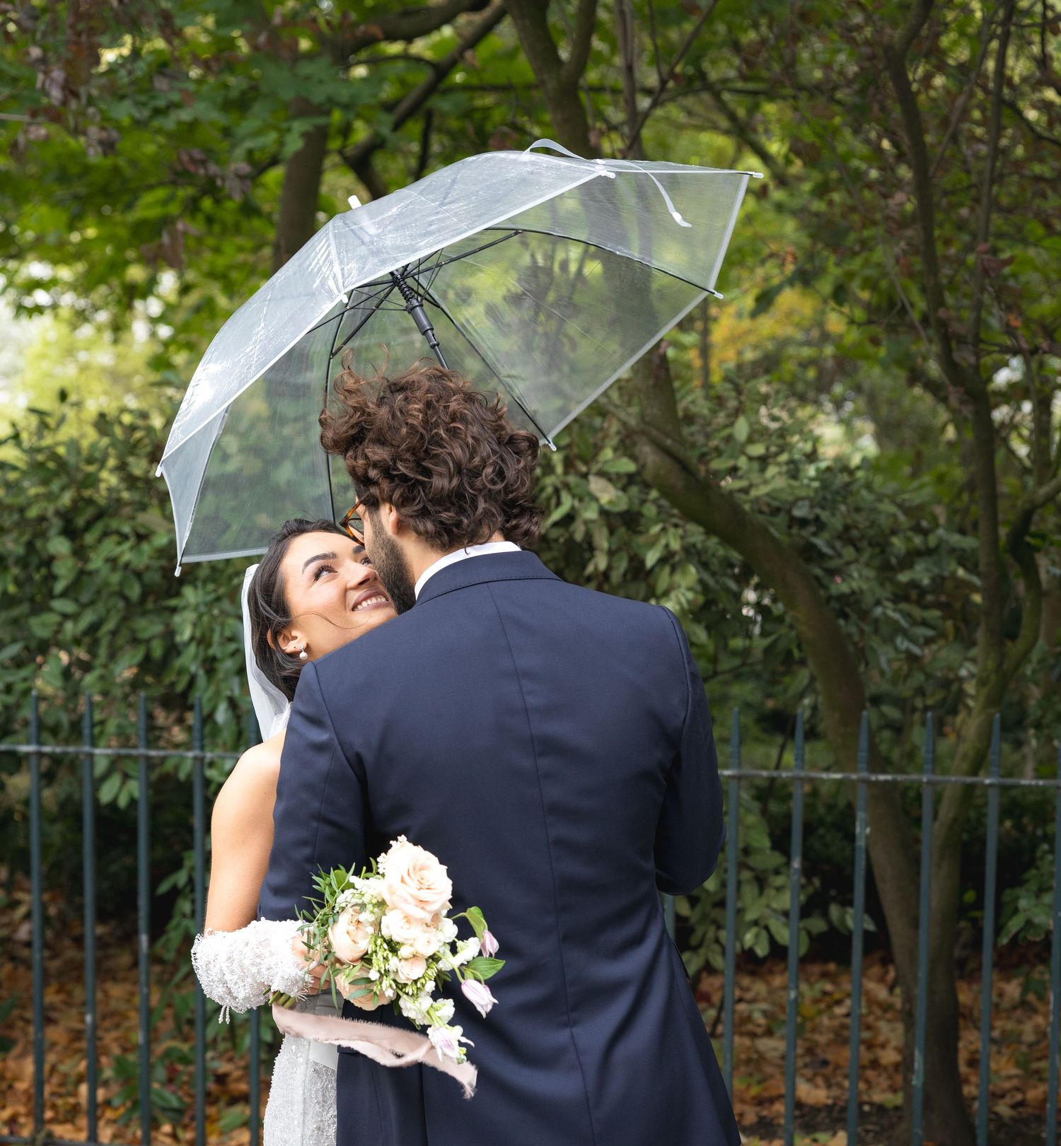 A couple shares an intimate moment under a clear umbrella in a garden setting.