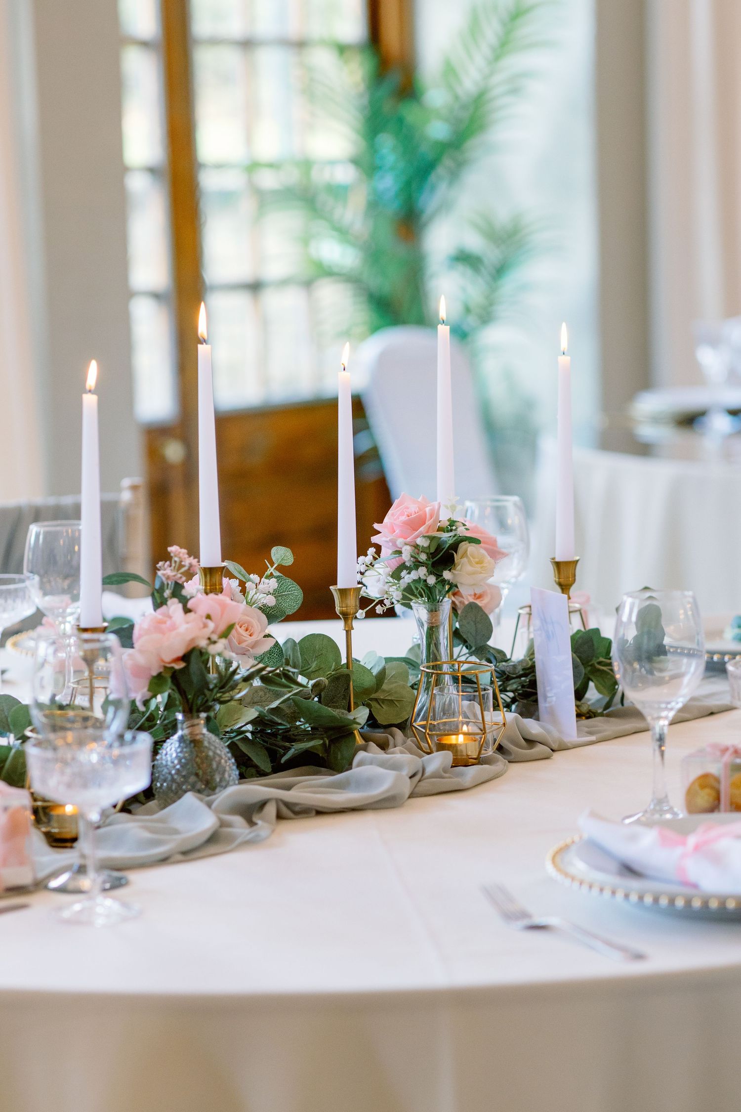 Elegant wedding reception table with white candles, pink flowers, and eucalyptus greenery in a bright sunlit venue.