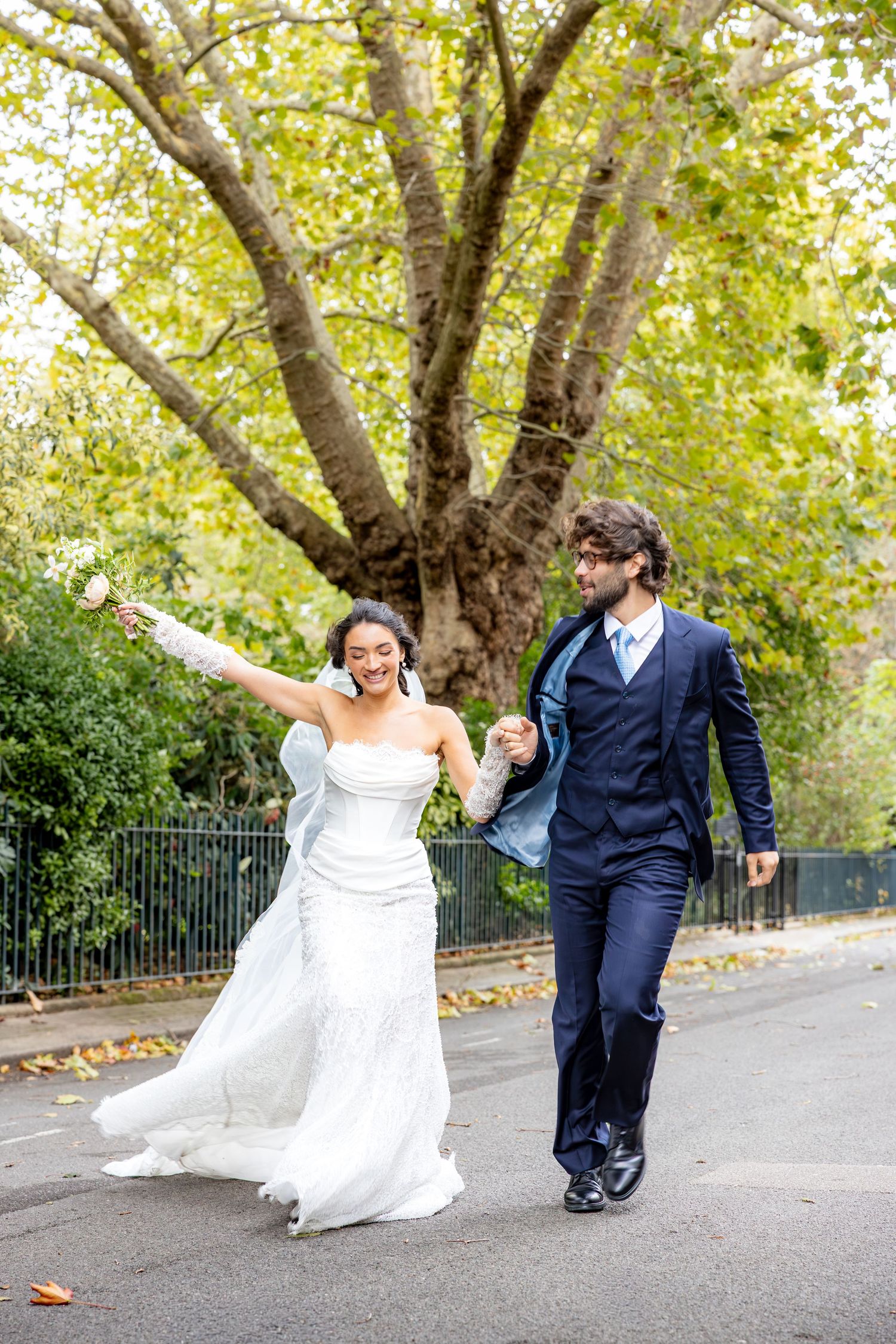 Playful bride and groom dance and spin together on a tree-lined path.
