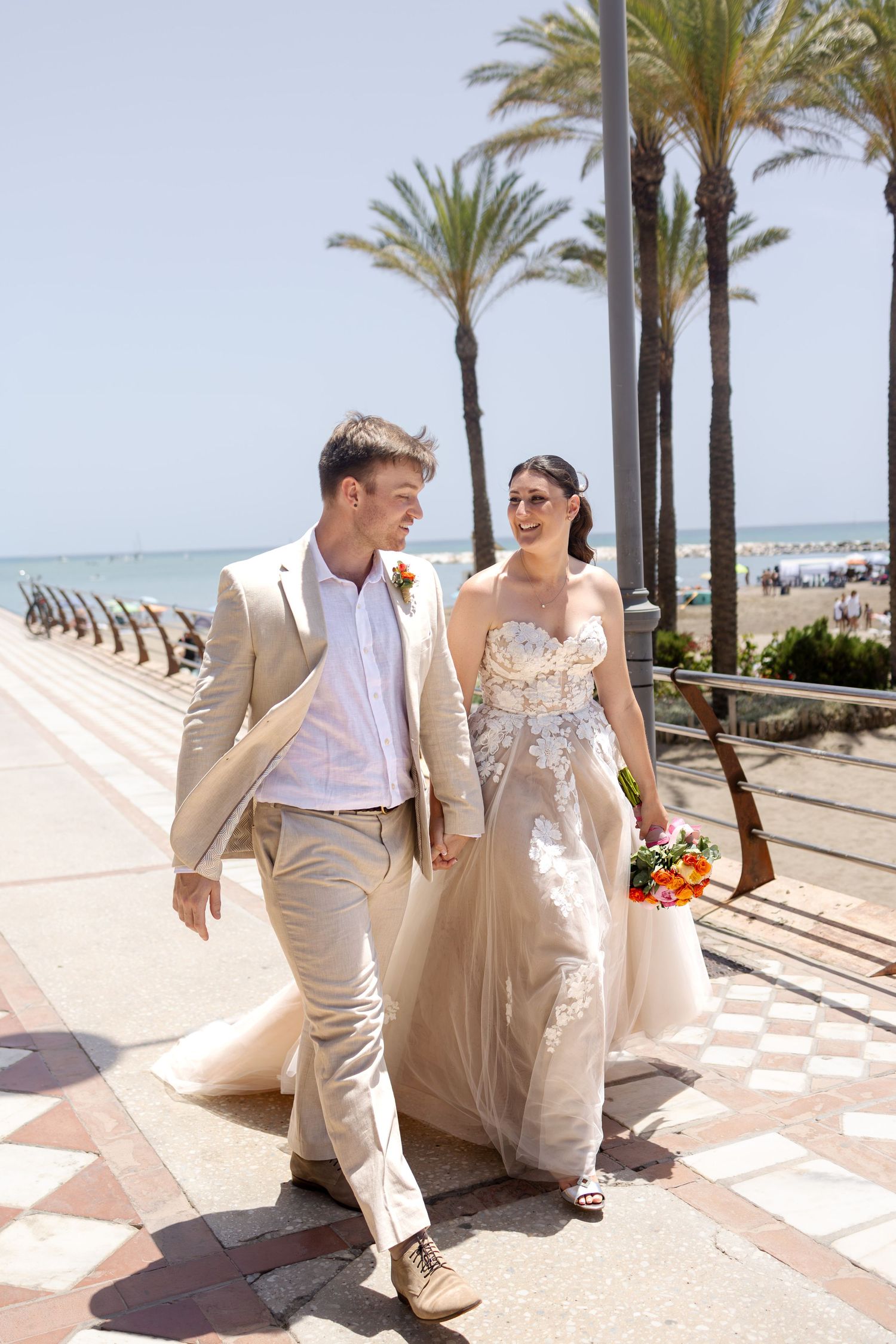 Bride and groom walk along sunny palm tree lined promenade near beach on wedding day.