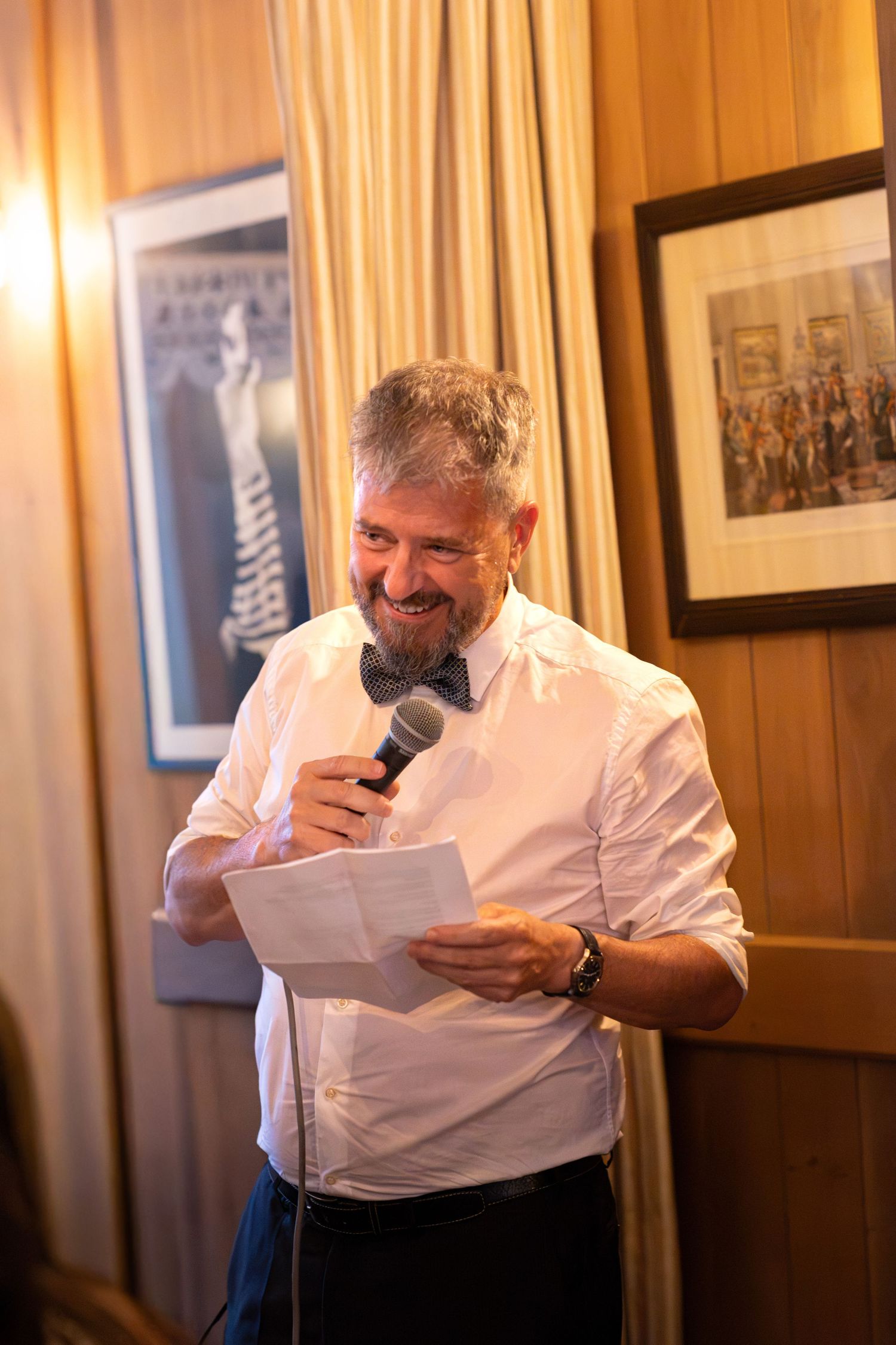 Smiling bearded man in white shirt and bow tie giving a speech at a wedding reception.