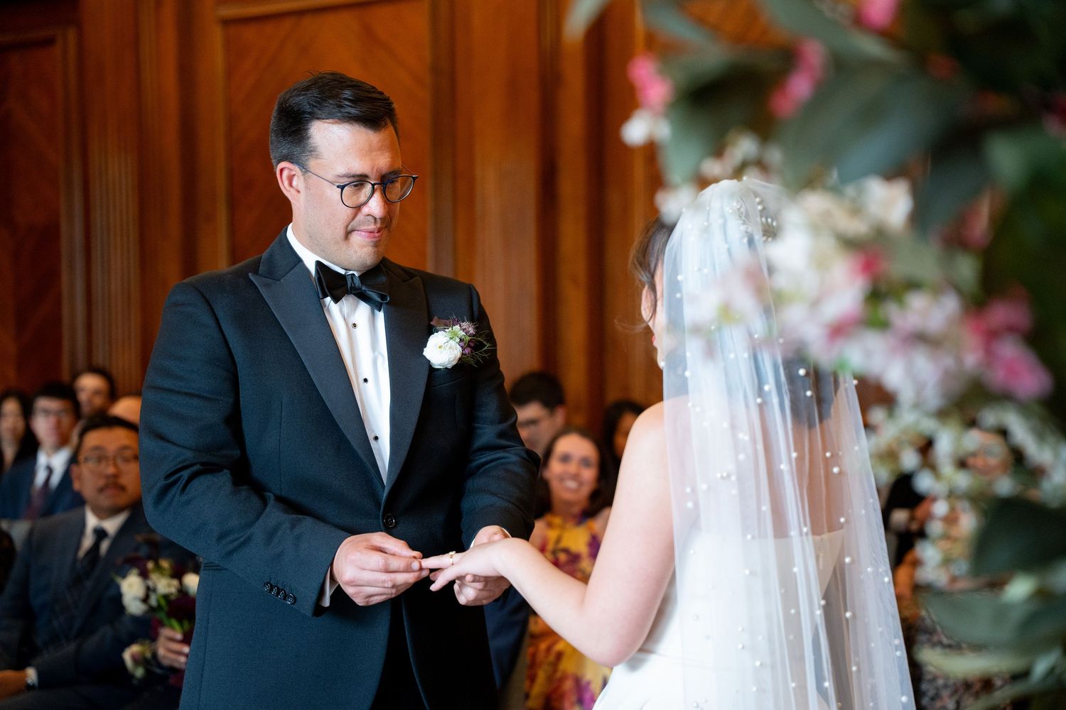 Groom in navy tuxedo placing wedding ring on brides finger during elegant ceremony with floral decor.